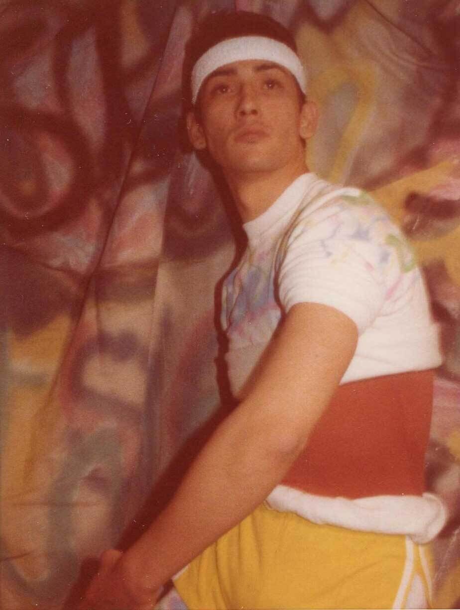 Young man in a white headband and colorful shirt poses in front of a graffiti-covered wall, wearing yellow shorts and a red waistband.