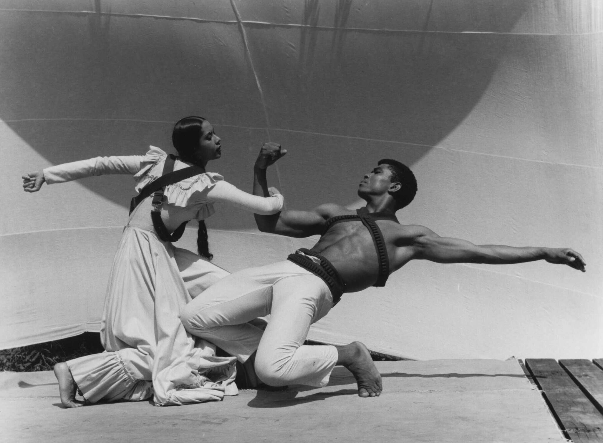 Two dancers perform a dramatic pose; the woman in a long dress and the man shirtless, both wearing harnesses, against a minimalist backdrop.