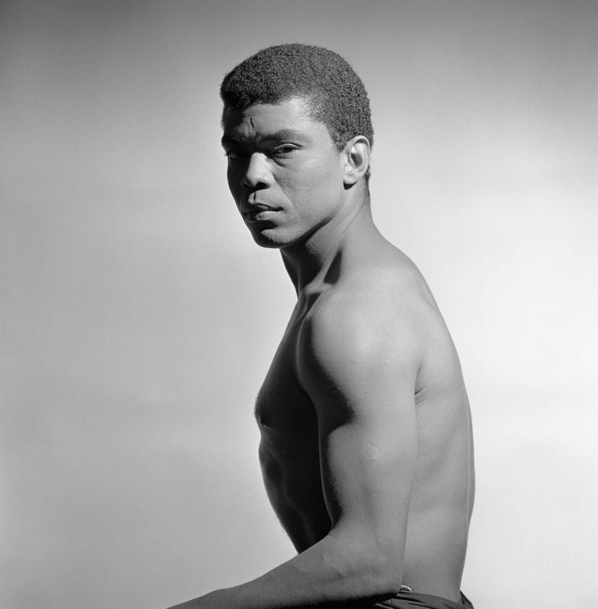 Black and white photo of a shirtless man with short hair, looking over his shoulder against a plain background.