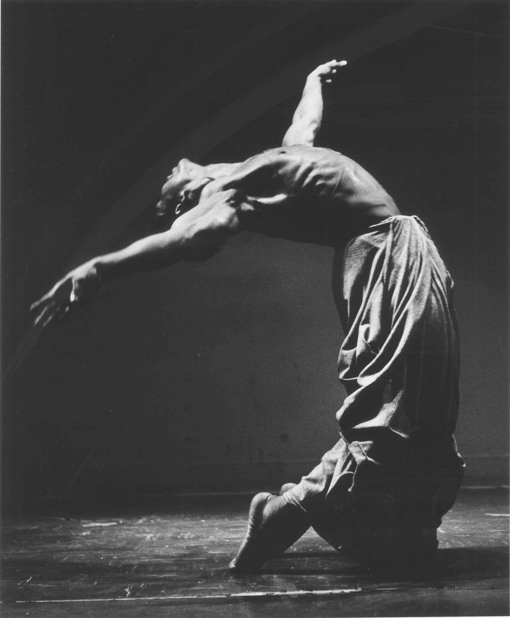 A dancer in loose pants arches backward with arms extended, captured in a dramatic black-and-white photograph.