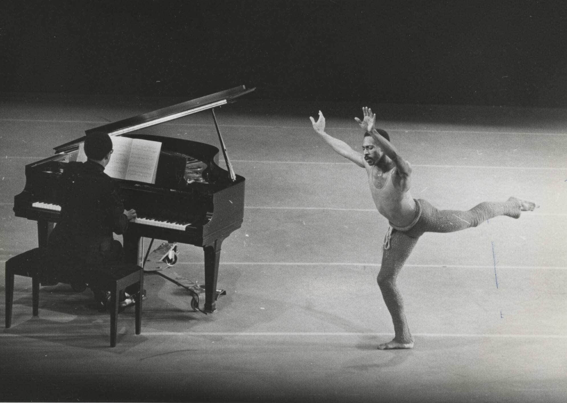 A pianist plays a grand piano while a dancer in a tank top and pants performs a ballet move on stage.