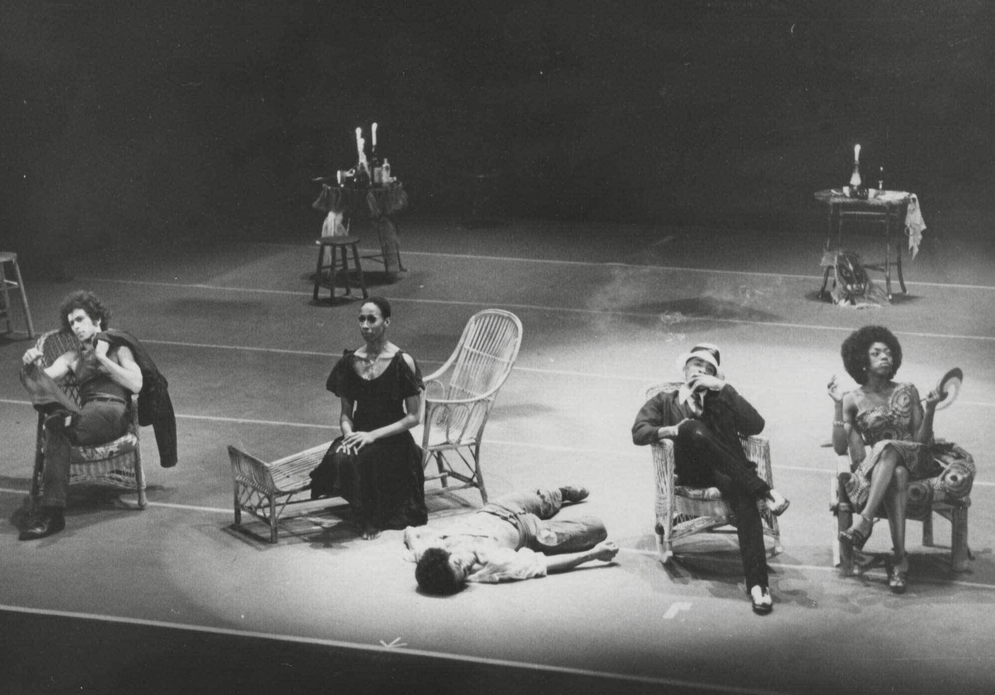 Four people sit in chairs on a stage, with one person lying on the floor. Tables with candles and bottles are in the background.