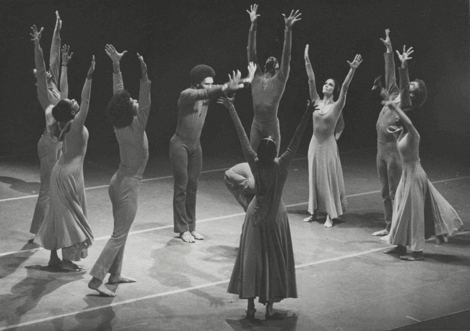 A group of dancers in flowing costumes and leotards perform on stage, raising their arms towards the sky in a circle formation.