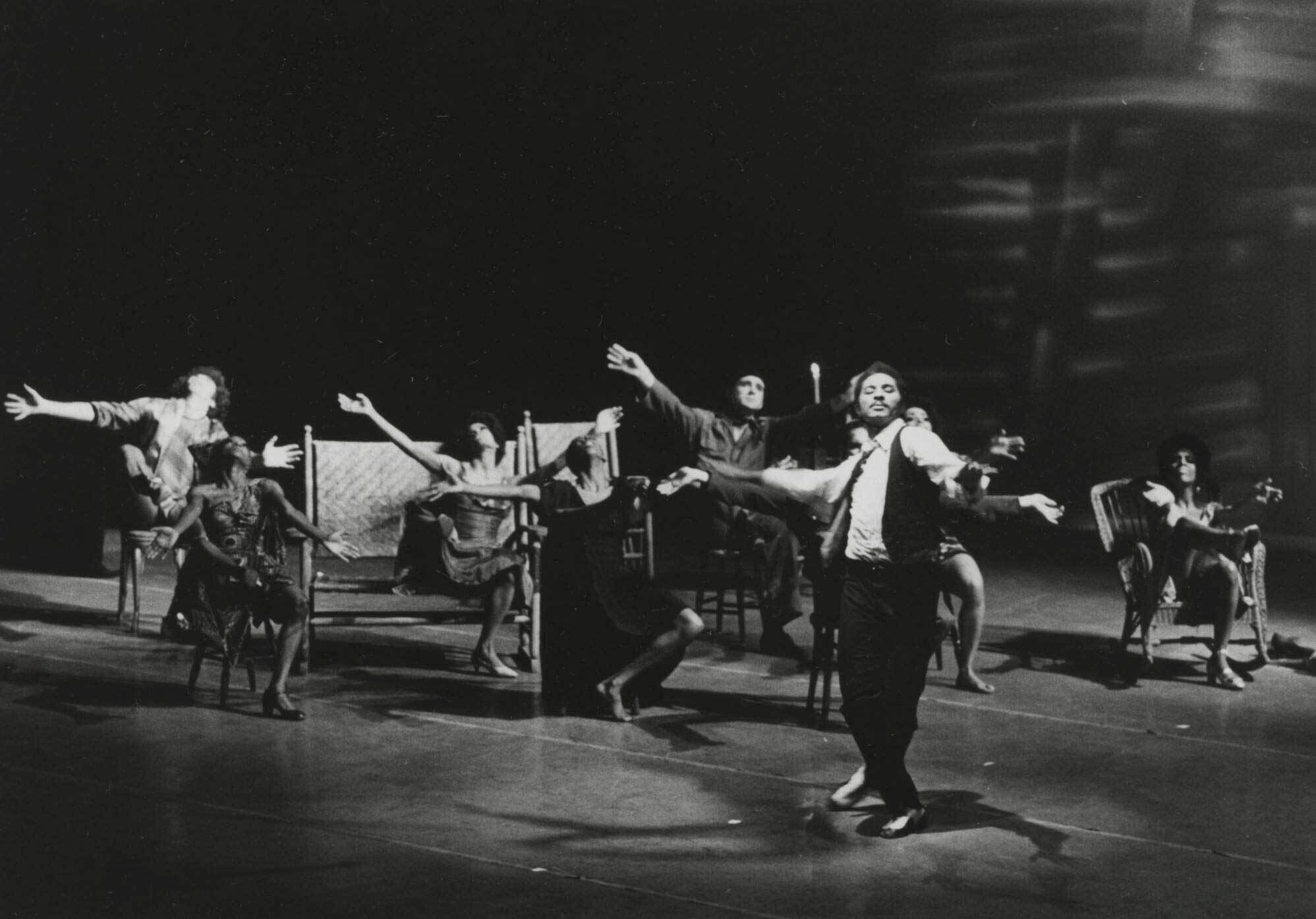 Performers on stage in dramatic poses, with arms outstretched and expressive movements, set against a dark background.