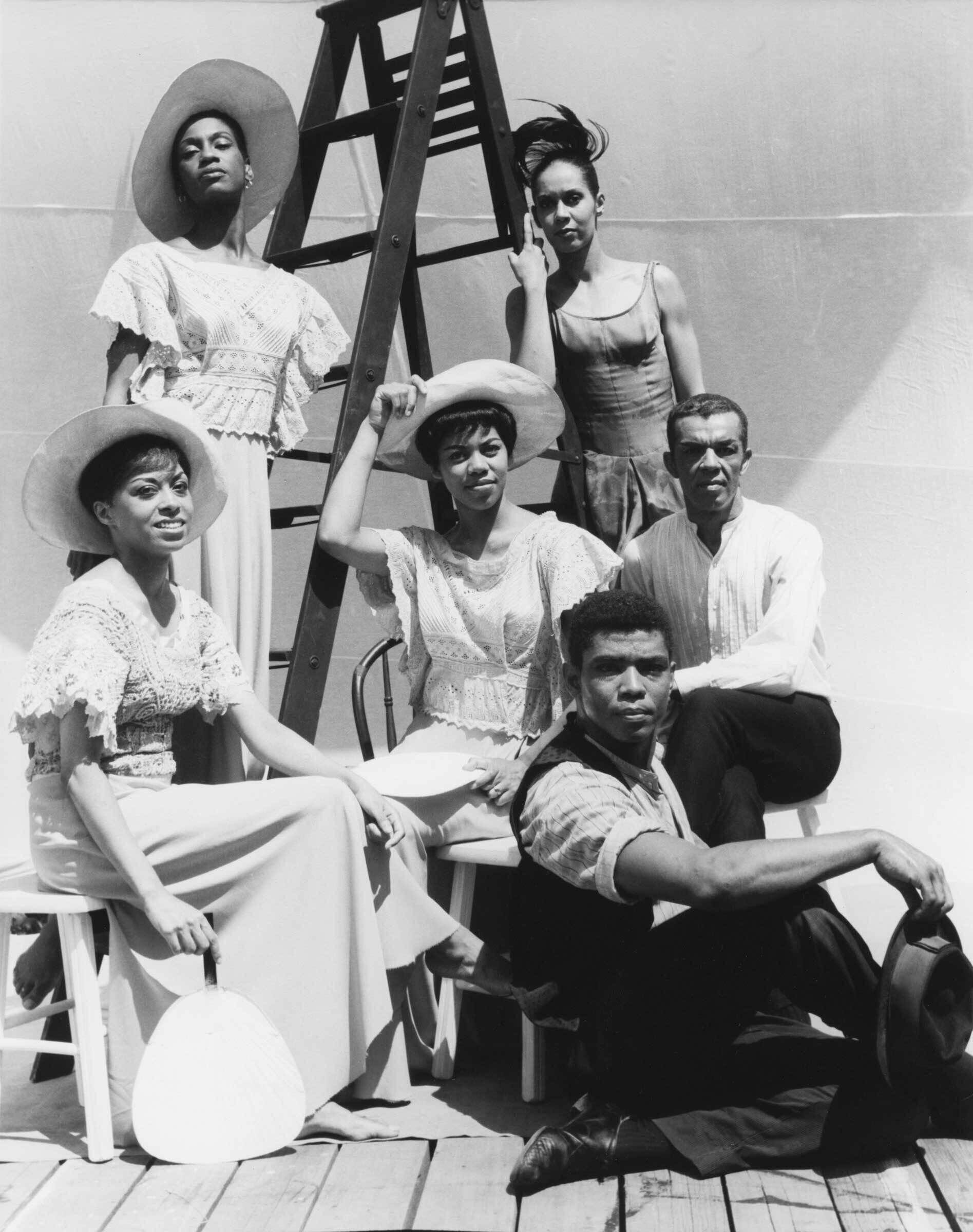 Six people pose around a ladder, with three women seated, two women standing, and one man sitting on the floor, all in vintage attire.