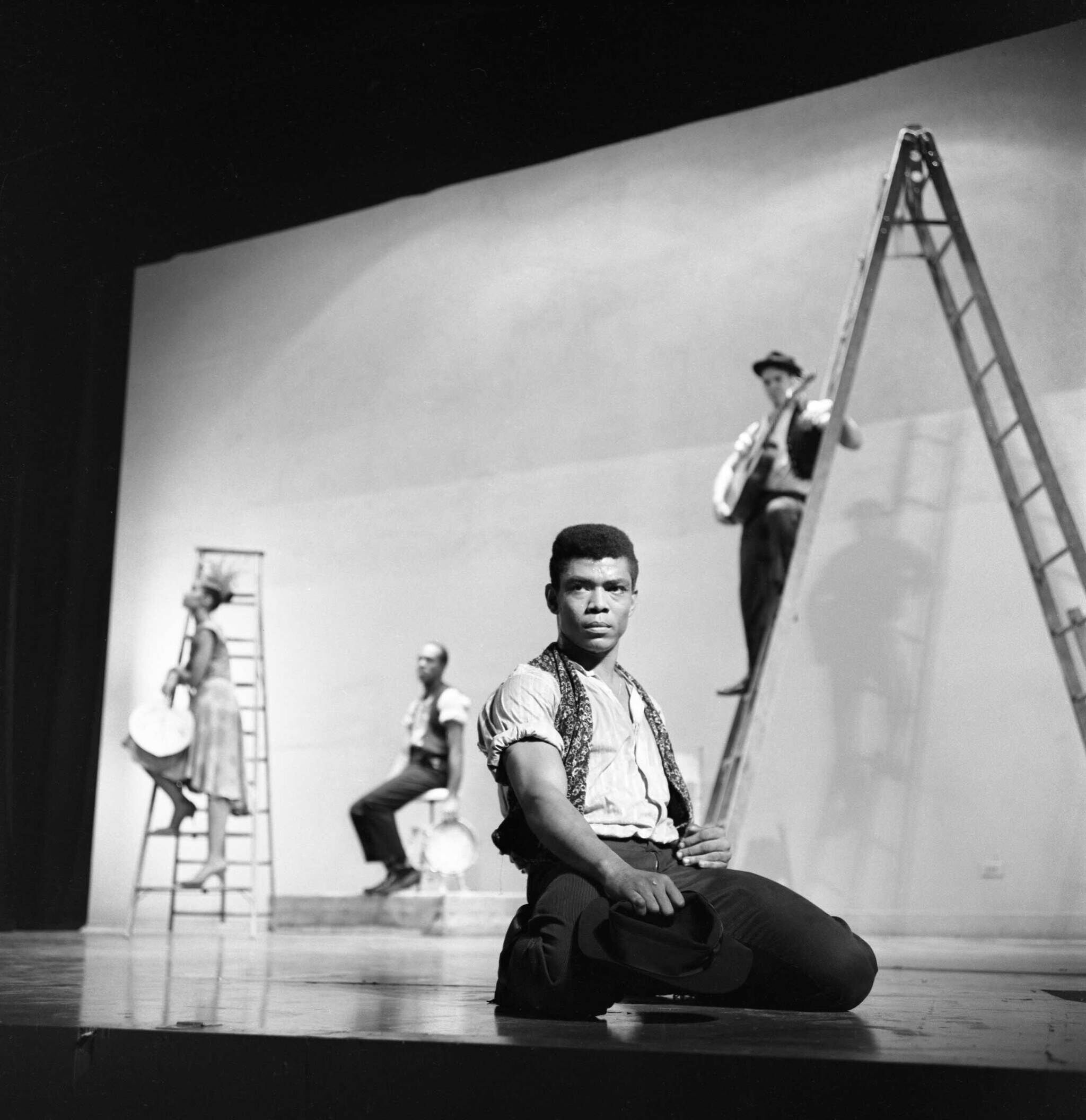 A man kneels on stage, holding a hat. Three other performers with instruments are on ladders in the background.