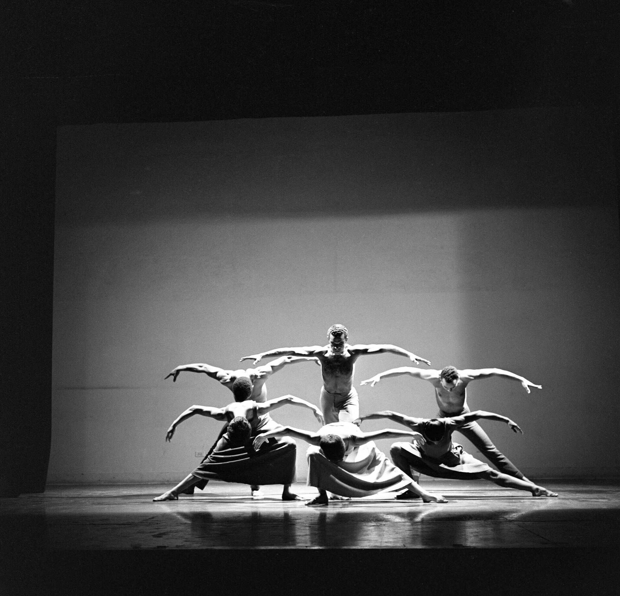 Five dancers in a dramatic pose on stage, arms outstretched, creating a symmetrical formation under spotlight in a black and white setting.