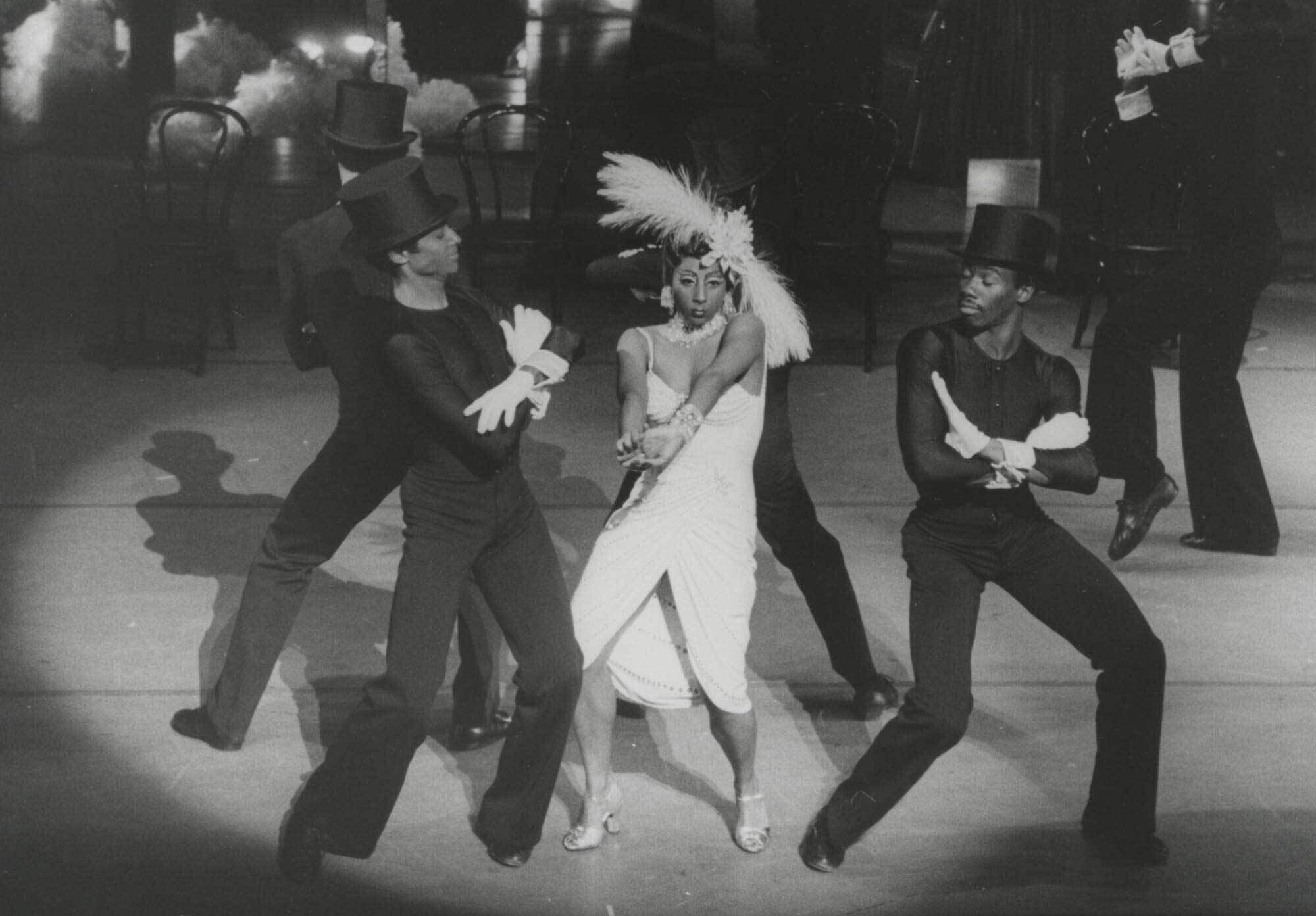 A woman in a feathered headdress and white dress dances with two men in top hats and gloves on stage, with chairs and lights in the background.