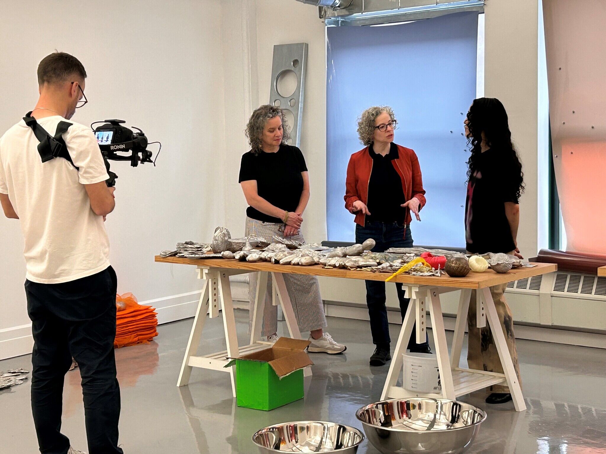 A person films three individuals discussing various objects on a table in a well-lit room.