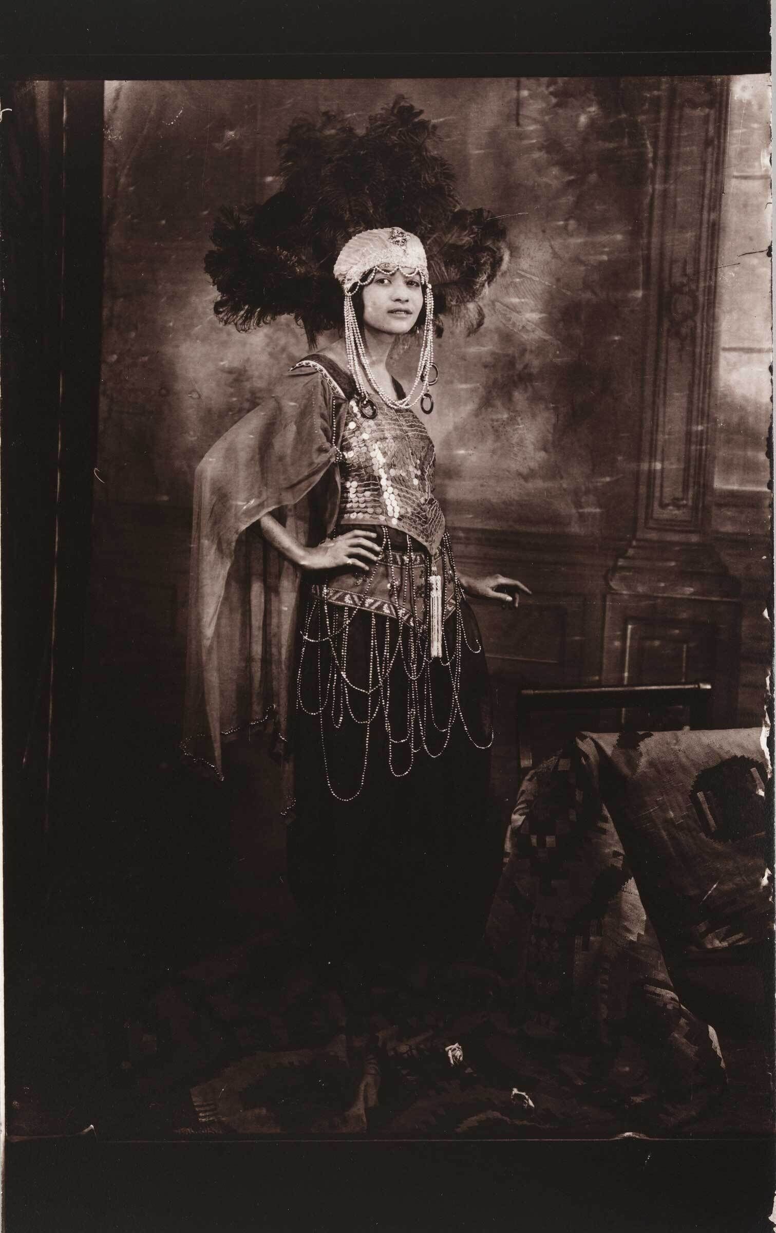 A person in an elaborate costume with a feathered headdress and sequined top, posing with one hand on their hip in a vintage, sepia-toned photograph.