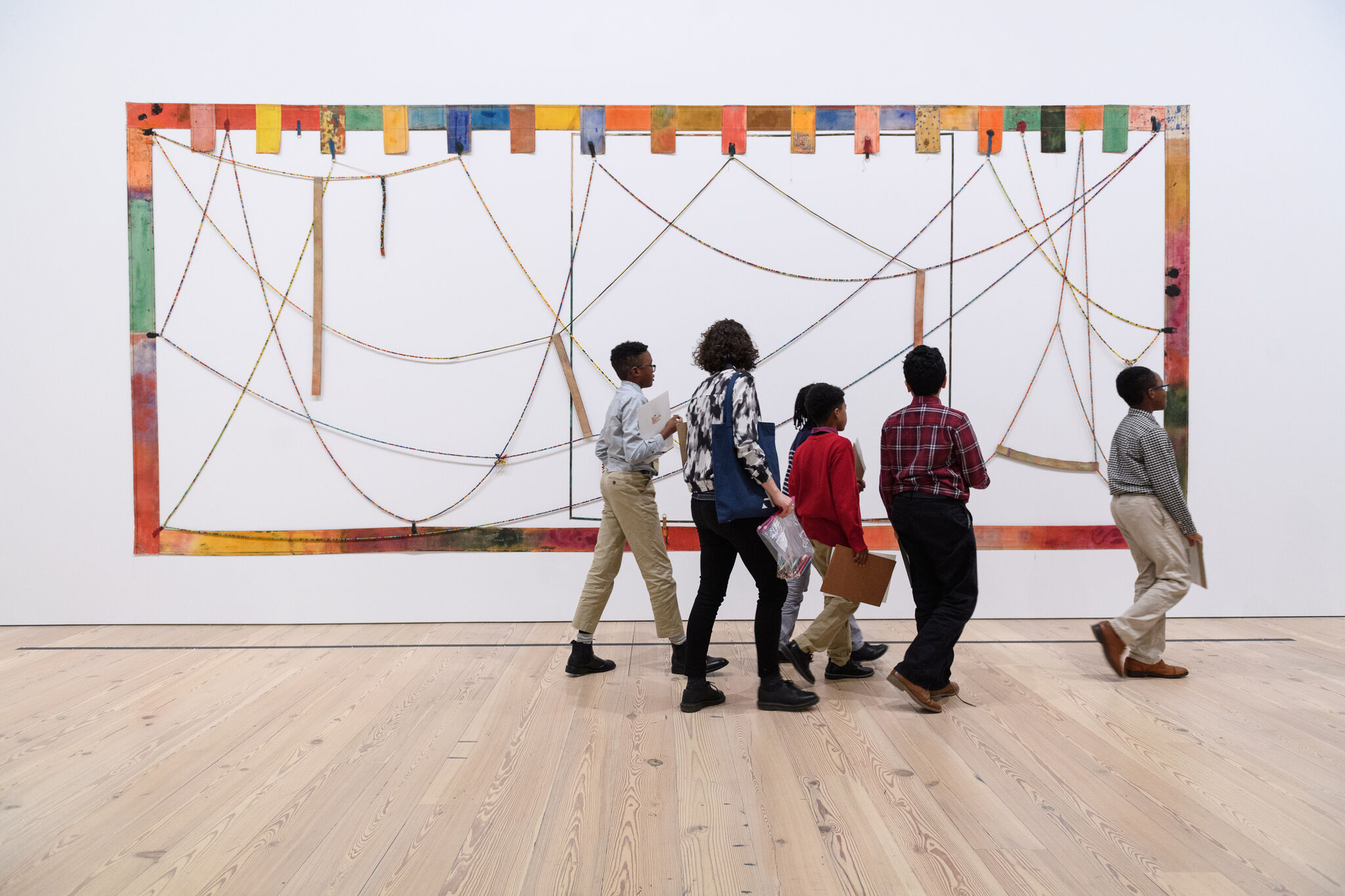 A group of people walks past a colorful abstract art piece with strings and rectangular shapes on a white wall in a gallery.