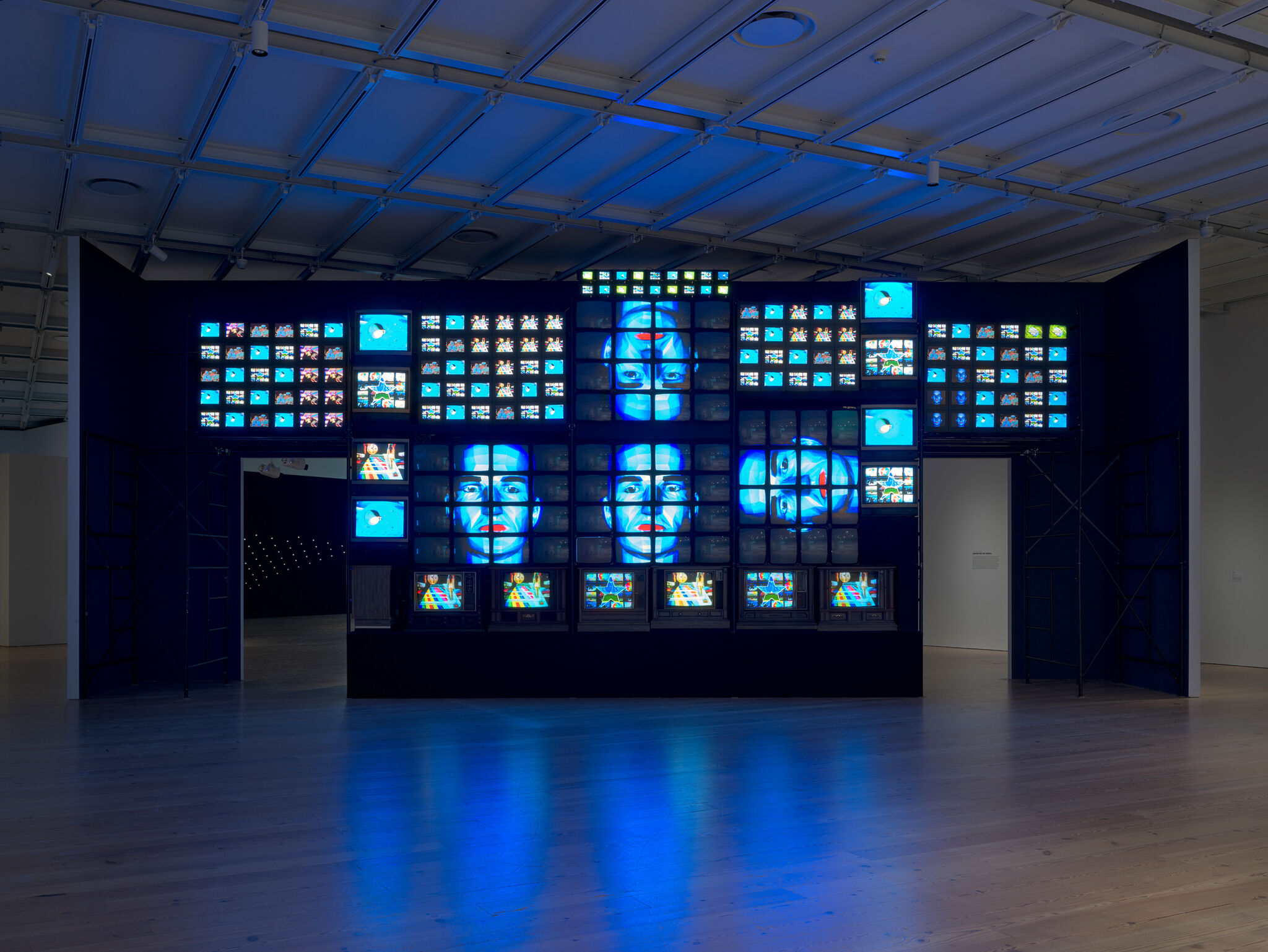 A large video wall installation in a gallery, displaying multiple screens with blue-toned images, including faces and abstract visuals. 