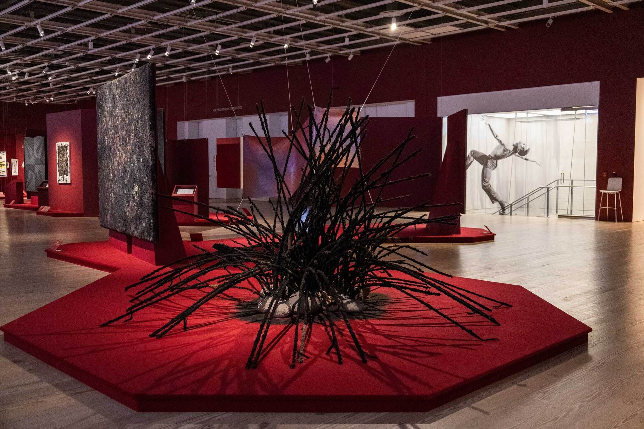 Modern art gallery with red platforms, abstract sculptures, and large paintings. A black-and-white photo of a person in motion is on the back wall.