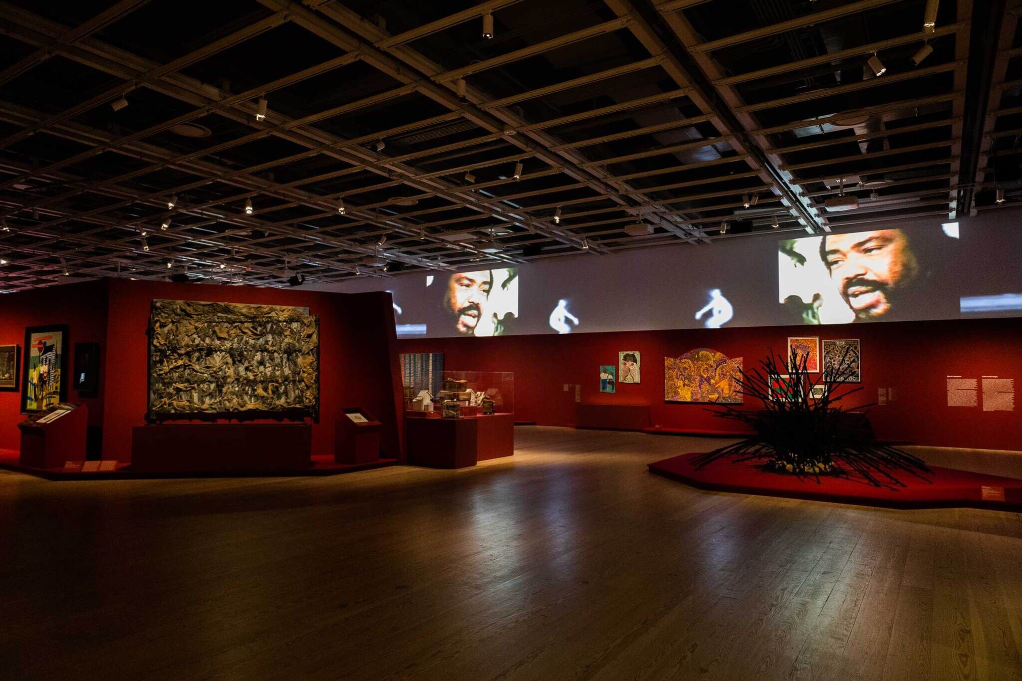 Art gallery with red walls, featuring various paintings, sculptures, and a large video projection of a man's face on the upper wall.