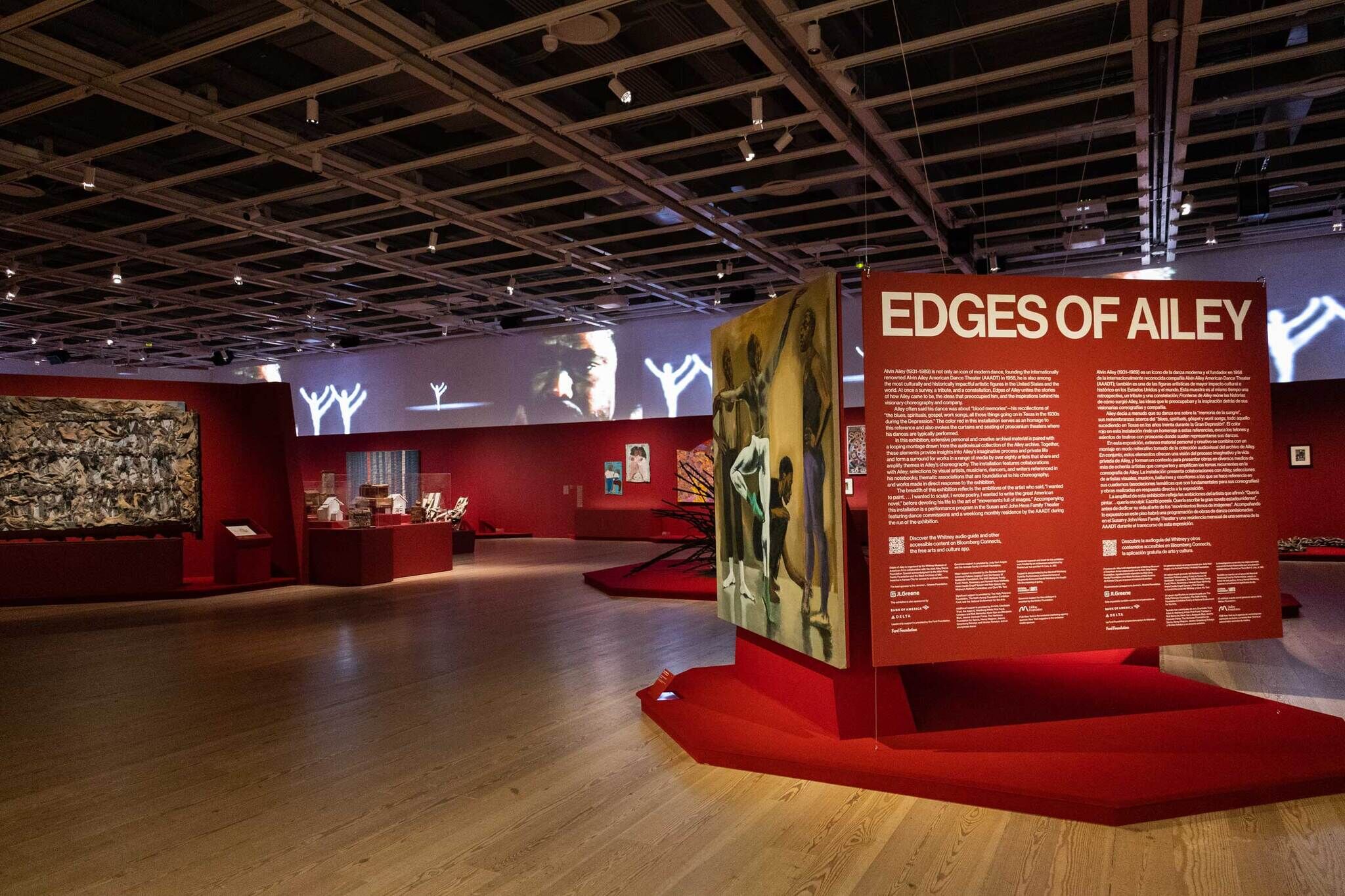 Museum exhibit titled "Edges of Ailey" with red walls, various artworks, and informational panels. Ceiling lights illuminate the spacious gallery.