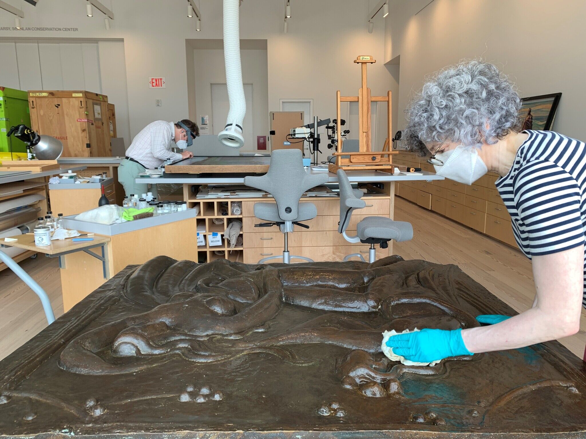 Two conservators work on art restoration in a well-lit studio. One cleans a large sculpture, while the other examines a piece on a table.