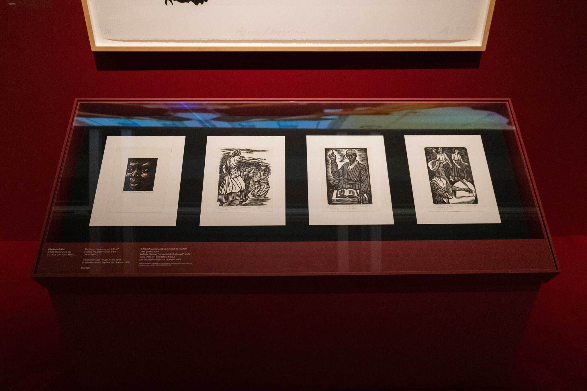 Four framed black-and-white prints displayed in a glass case against a red background, each depicting different scenes and figures.