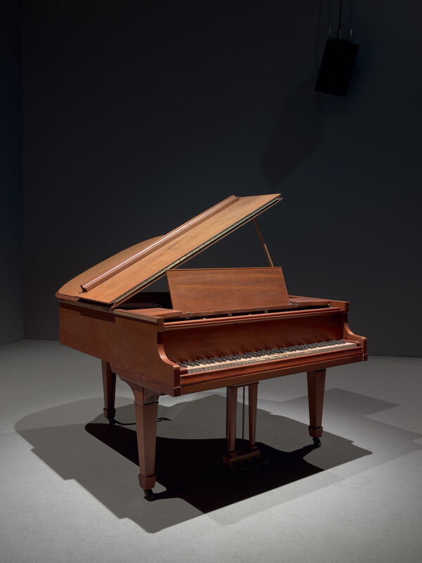 A wooden grand piano with its lid partially open, placed in a dimly lit room with a dark background.