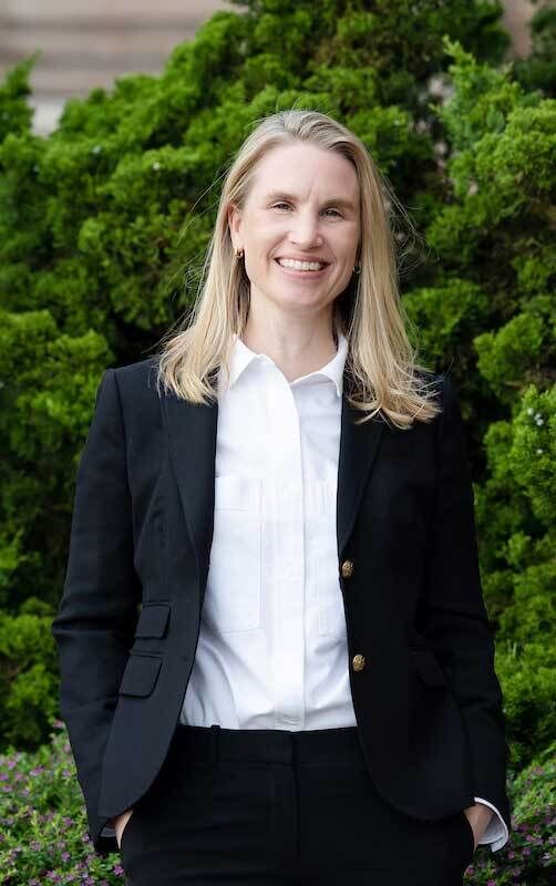 Smiling woman with blonde hair in a black blazer and white shirt stands in front of green foliage.