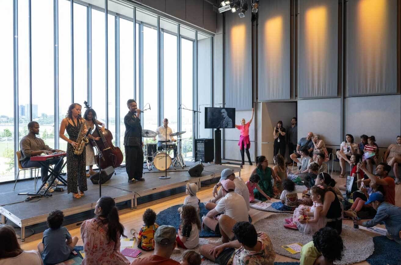 A live jazz performance by Jazz at Lincoln Center musicians at the Whitney Museum. The band, featuring a saxophonist, bassist, pianist, drummer, and vocalist, performs on a stage in front of a large glass window overlooking the city. An audience of families, including children, sits on the floor, enjoying the music in an intimate and relaxed setting.