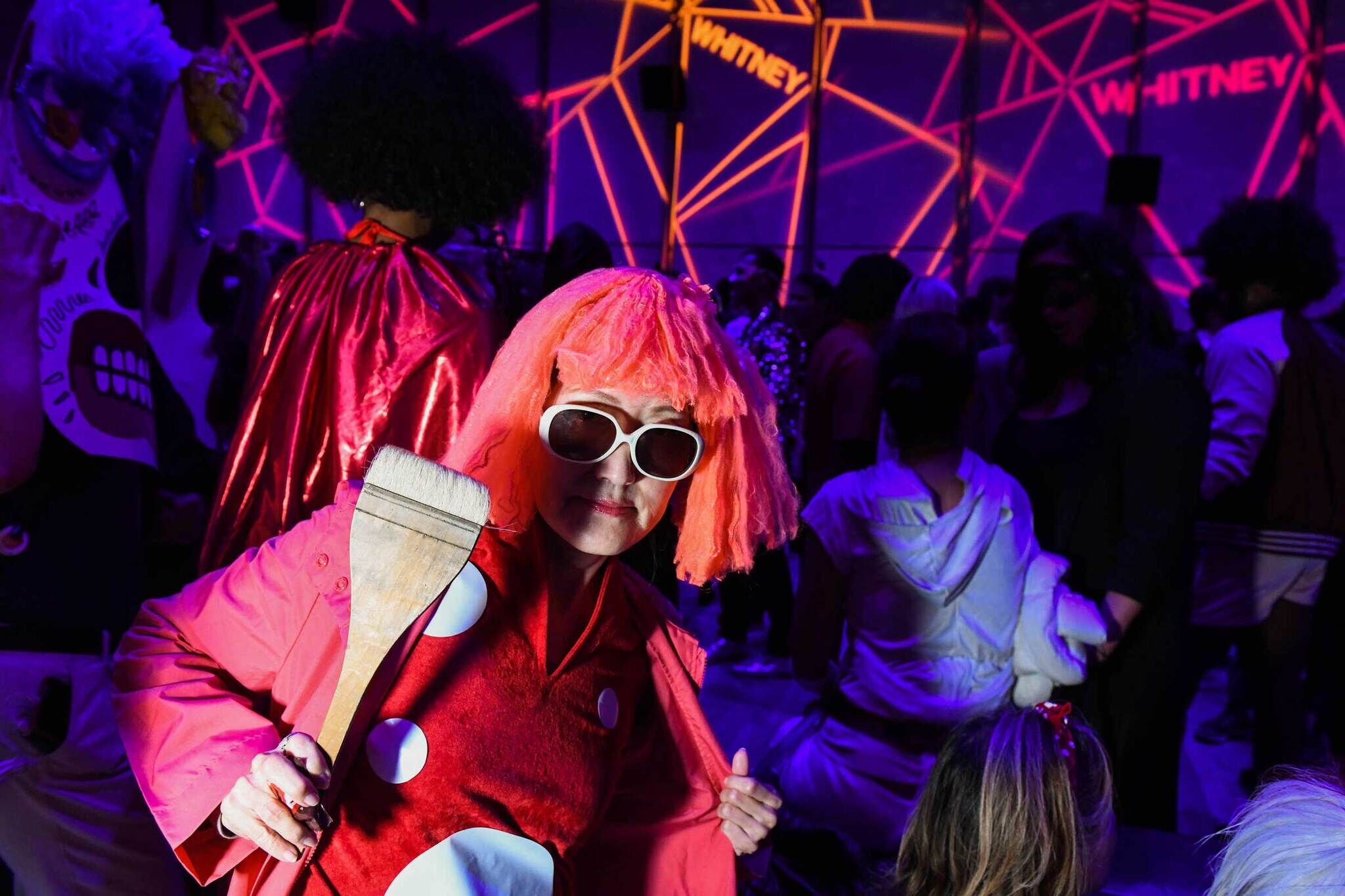 Person in a red costume with a pink wig and white sunglasses holds a large paintbrush at a lively event with neon lights and "Whitney" on the wall.