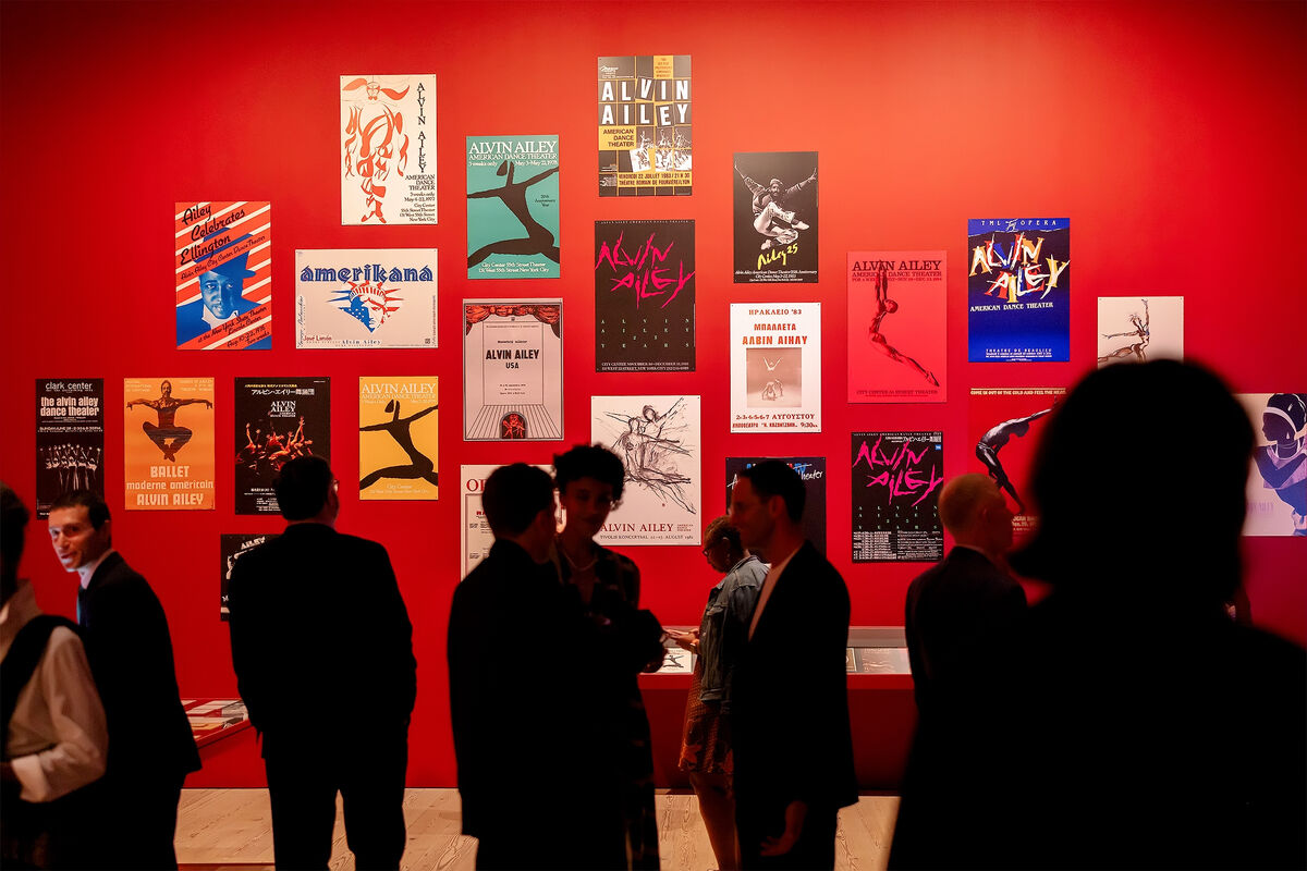 People viewing a red wall adorned with various Alvin Ailey posters at an art exhibit.