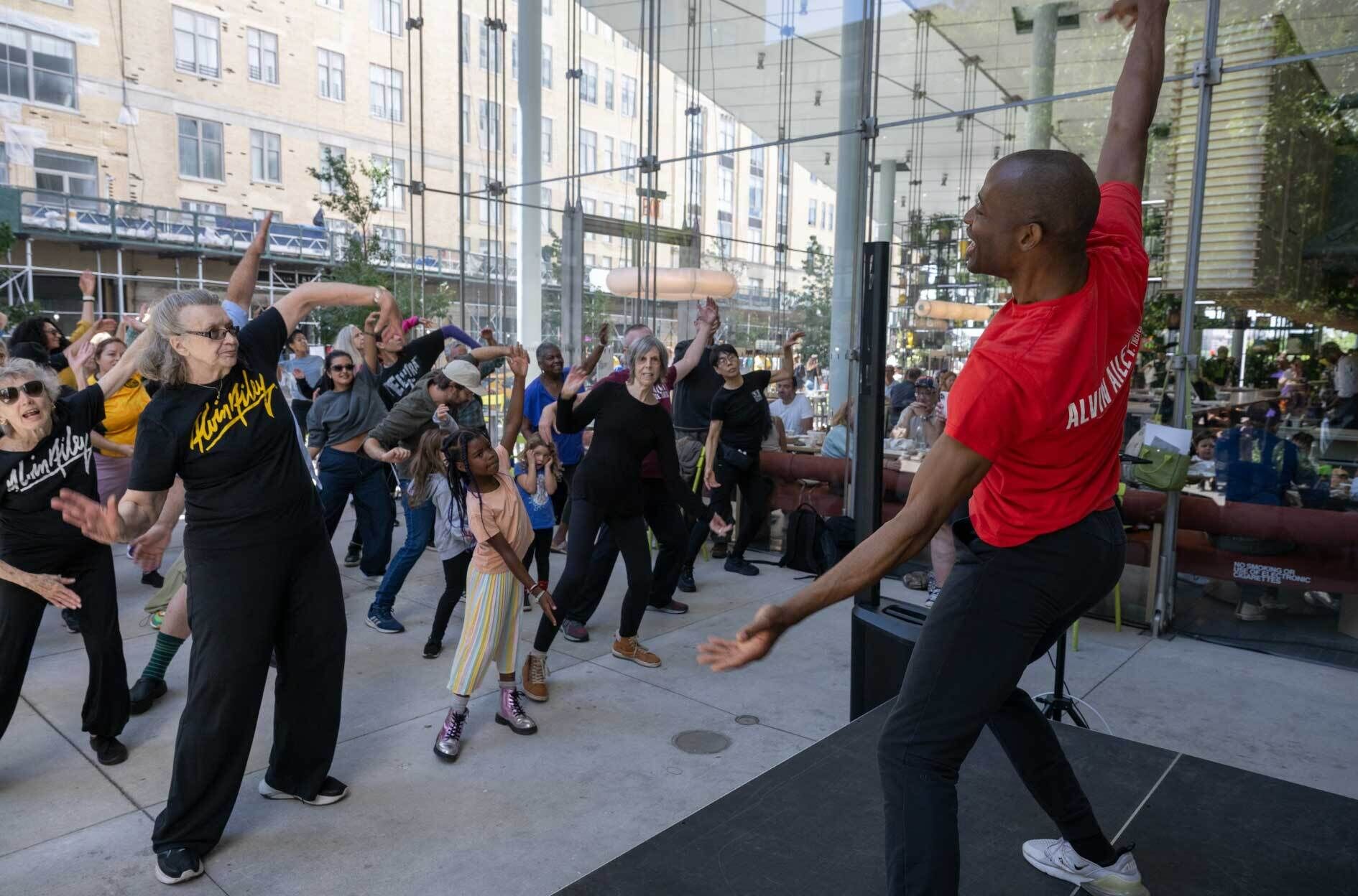  A dance class at the Whitney Museum led by an instructor from Ailey Extension. The instructor, wearing a red Alvin Ailey t-shirt, energetically leads a diverse group of participants, including children and seniors. The participants follow his movements with enthusiasm in an outdoor space with large windows, while onlookers can be seen in the background enjoying the lively atmosphere.