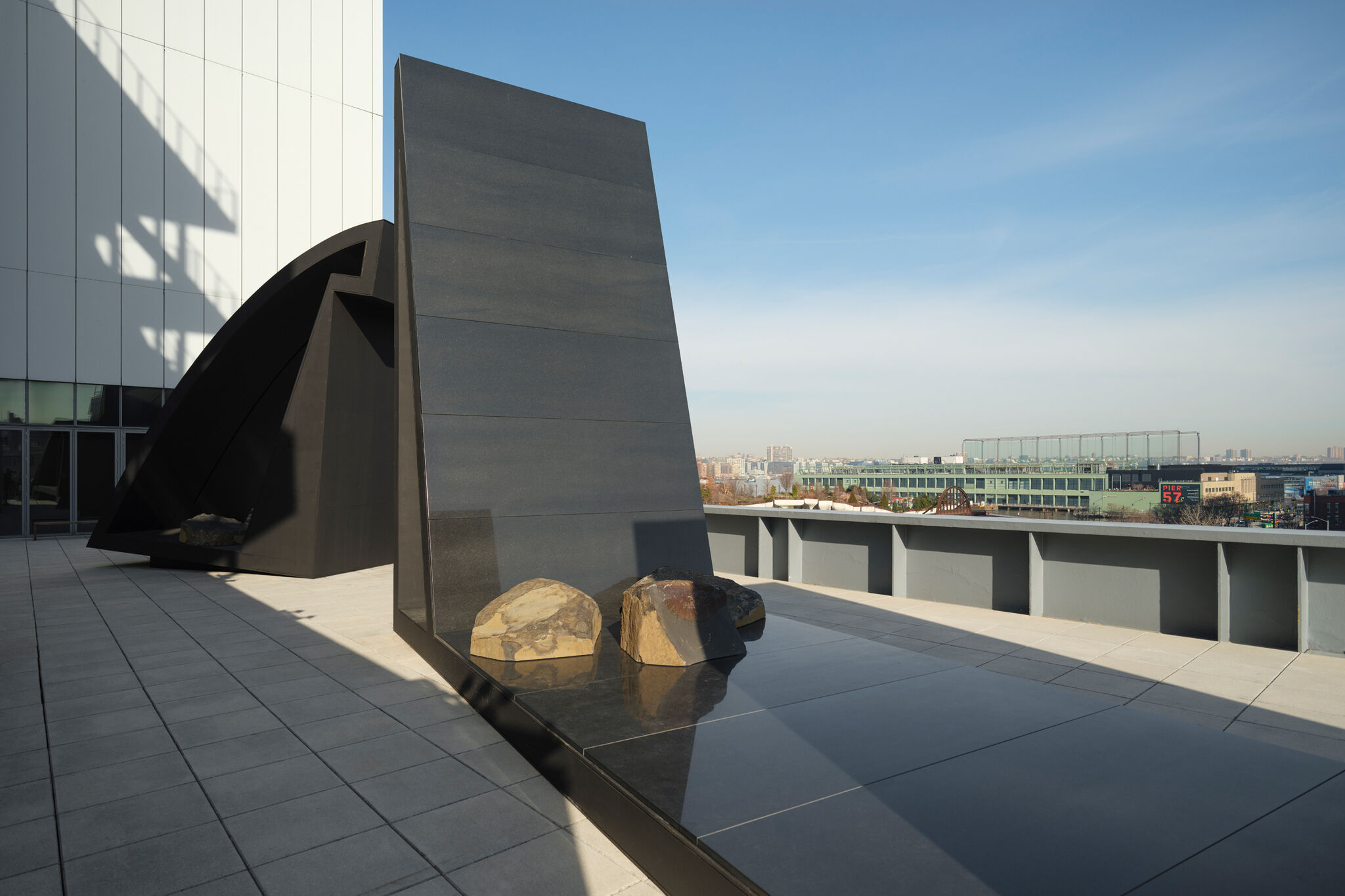 Modern rooftop sculpture with large black panels and rocks, set against a cityscape under a clear blue sky.