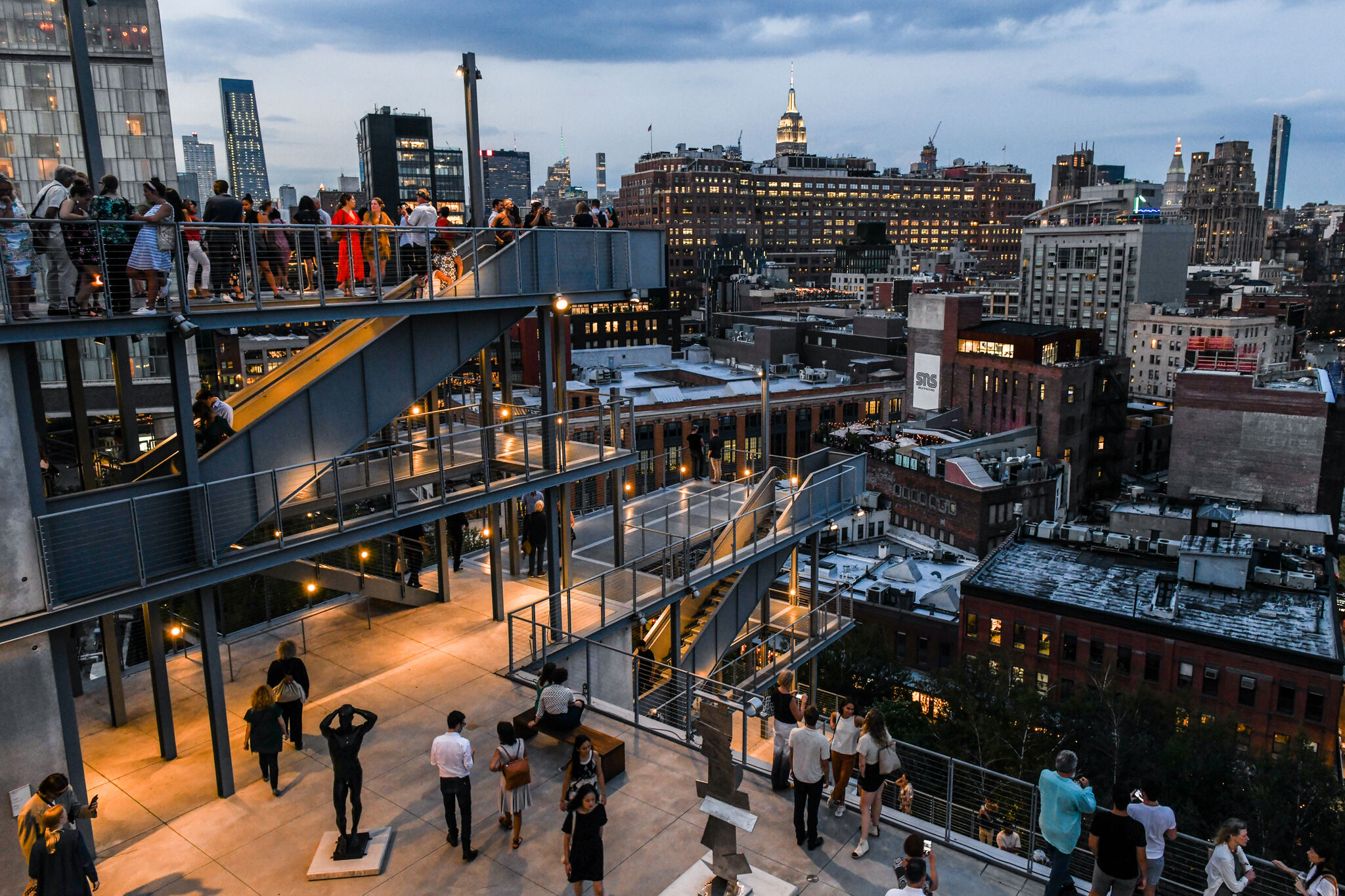 People gather on a multi-level outdoor terrace overlooking a cityscape at dusk, with buildings and a lit skyline in the background.