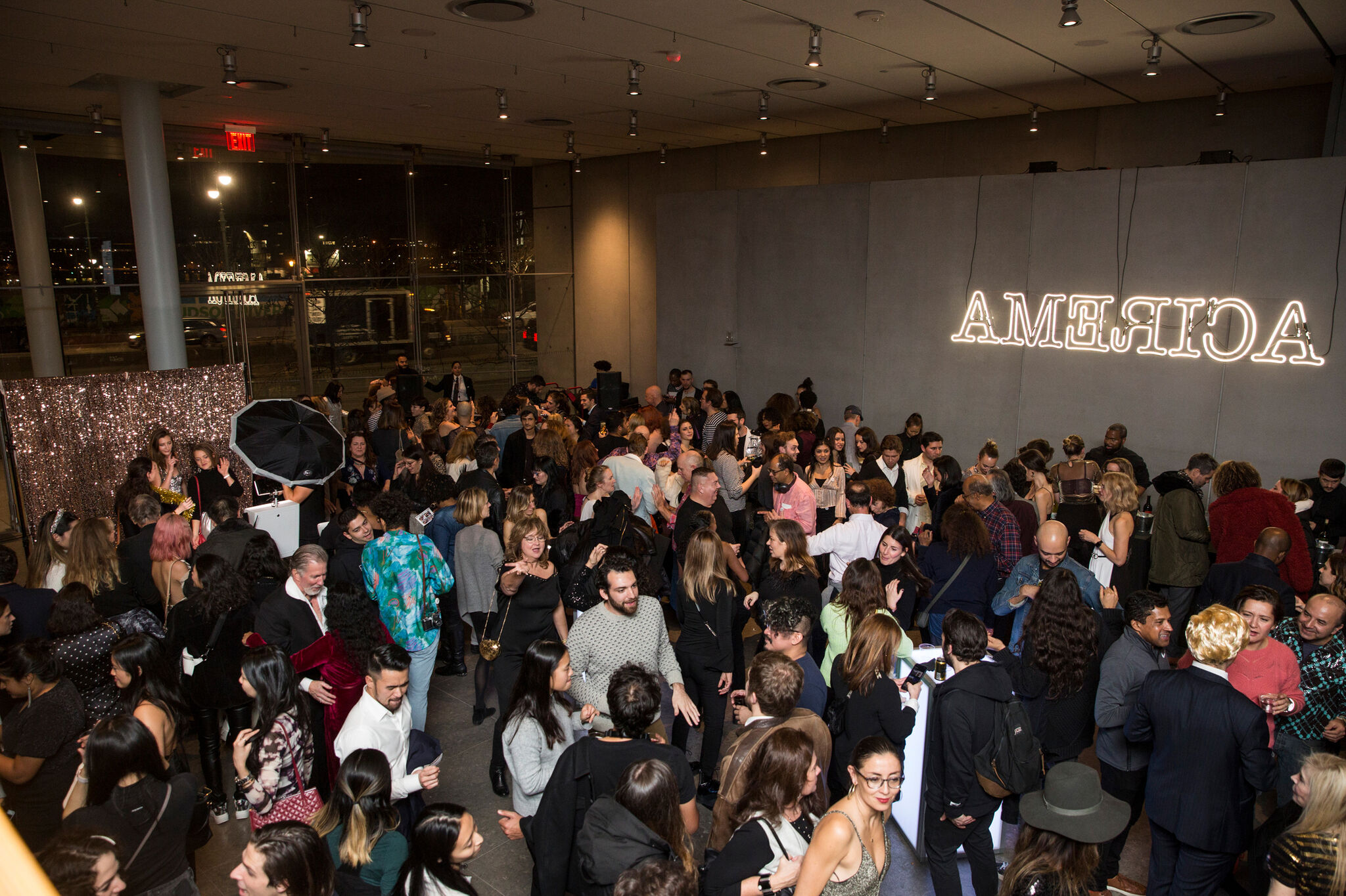 The photograph shows a large gathering of people in a dimly lit indoor space, at a party standing, mingling, and chatting with one another. The background features a large sign with the word "AMERICA" illuminated in bright, white neon lights, but the letters are reversed, creating a mirrored effect. In the foreground, people are dressed in a variety of styles, from casual to formal attire. The space has high ceilings and large windows, allowing a view of the outside night scene. There is a shiny, sequined backdrop in one corner, where some individuals appear to be posing for photos, with a professional camera setup nearby.