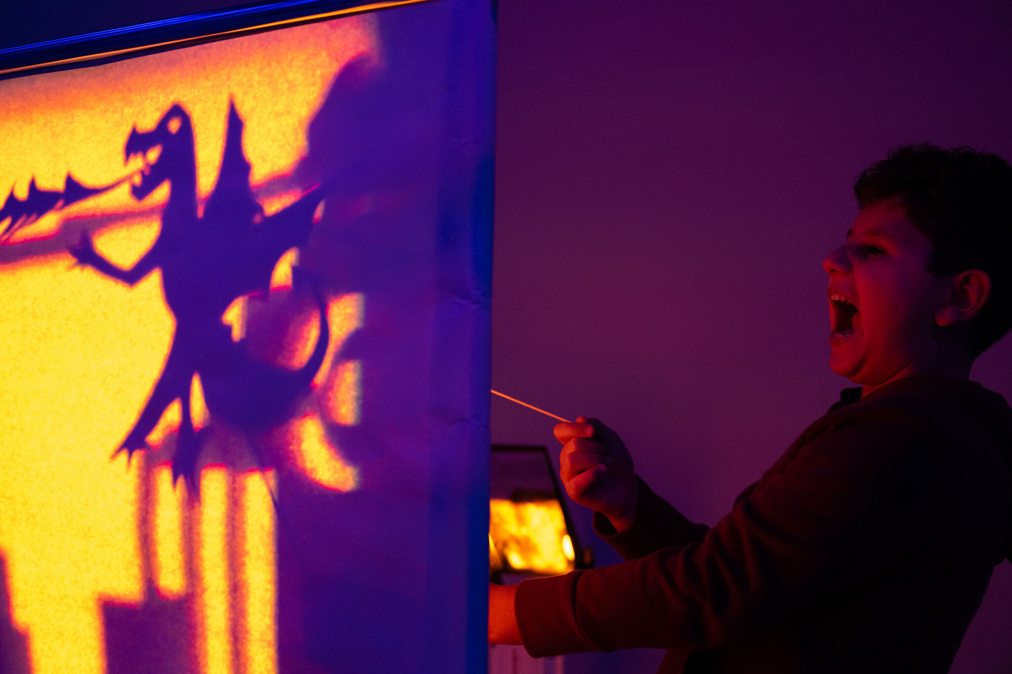 A child enthusiastically holds up a stick controlling a dragon shadow puppet, which casts a large, vivid silhouette onto a brightly lit surface. The child appears to be roaring playfully, mimicking the dragon's pose, while the scene is illuminated with dramatic, colorful lighting, creating a vibrant and imaginative atmosphere.