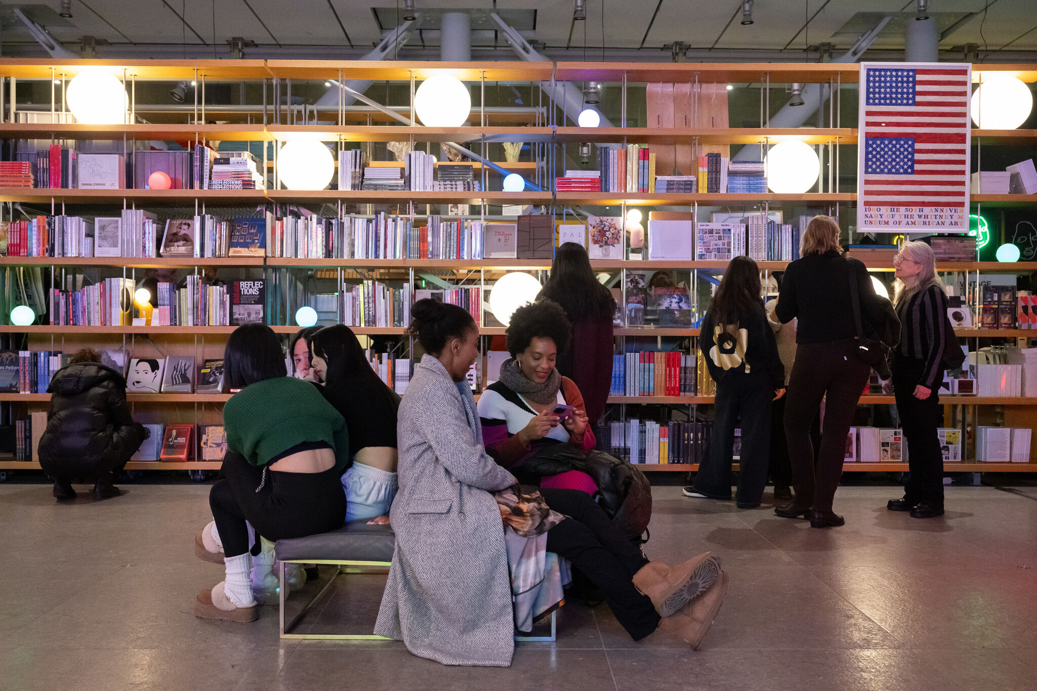 The photograph shows a bookstore with a group of people engaging in various activities surrounded by shelves filled with books and decorative items. At the center, there are several individuals dressed in winter attire that are sitting on benches, interacting with each other, and using their phones. In the background, more people are standing near the shelves, browsing through the books. The shelves are neatly organized, with a mix of colorful books and modern decor, including glowing orb-like lights. On the right side, a large artwork or poster of the American flag is visible.