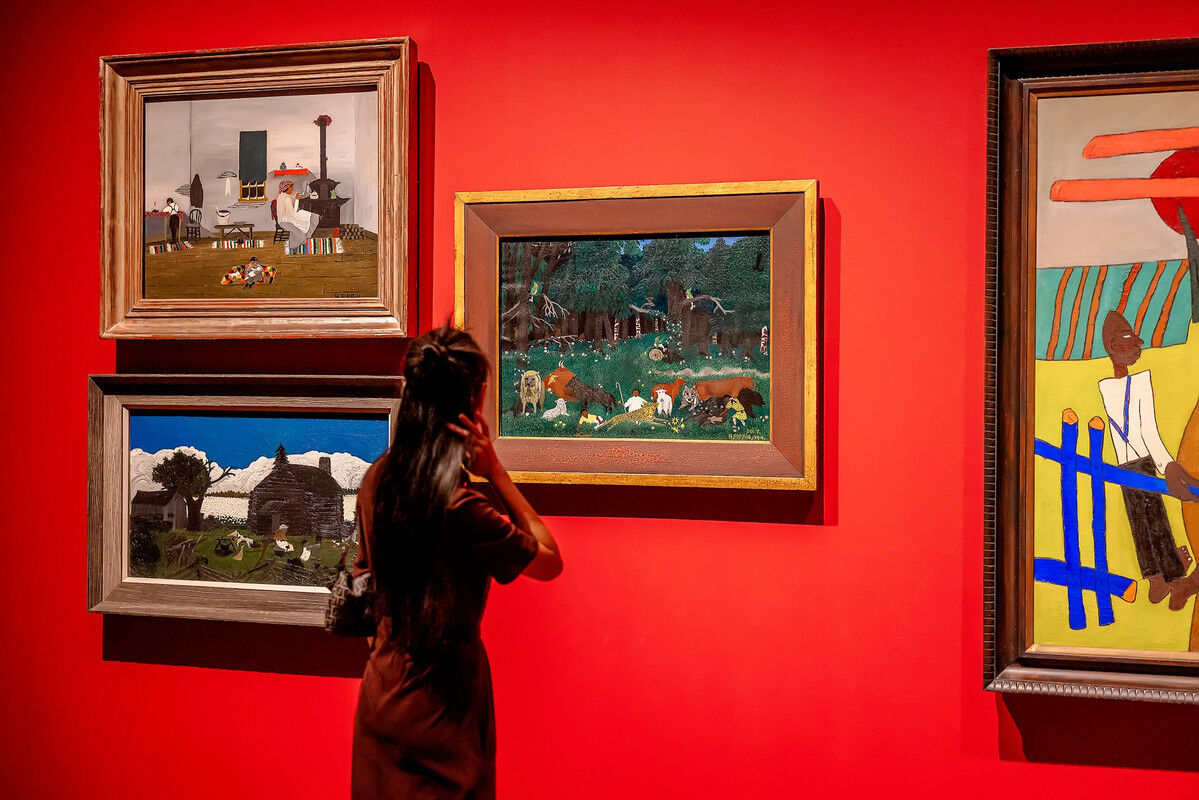 The photograph shows a woman with long dark hair, wearing a brown outfit,   standing in front of a brightly colored red wall, looking at several paintings on display. The paintings are framed in various styles, with some larger than others.  The artworks themselves include:  a scene on a farm with two people seated, another depicts three people in a home, and there is one that shows a scene of nature with animals in a wooded environment.