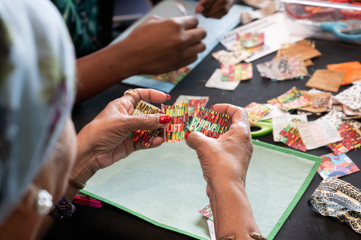 A seated woman is holding colorful paper cut-outs; a blank piece of paper and a spread of art materials can be seen on the table in front of her.