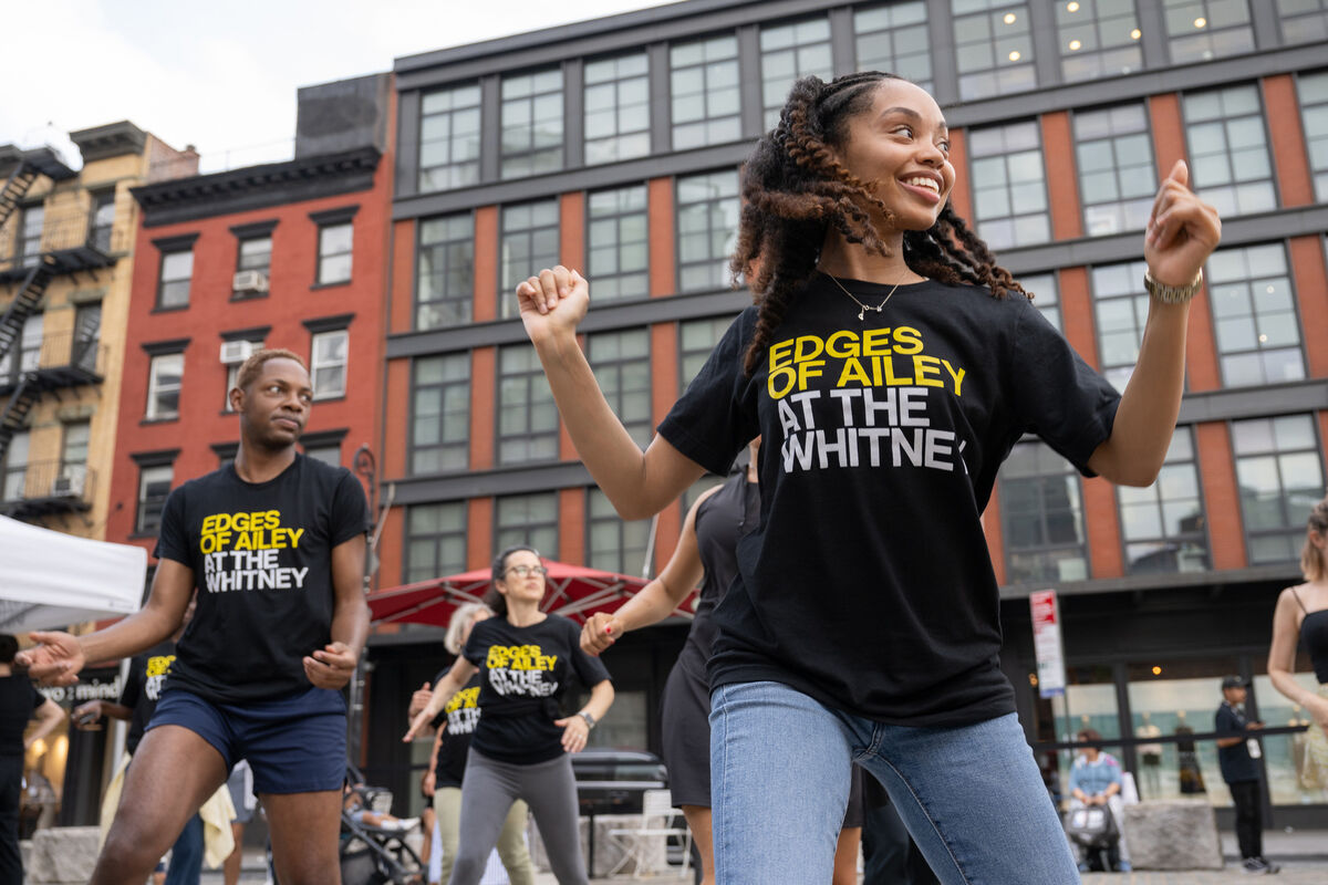 A dance class at the Whitney Museum led by an instructor from Ailey Extension. A diverse group of participants, wearing matching shirts with the text "Edges of Ailey at the Whitney" dance with enthusiasm in an outdoor space. 