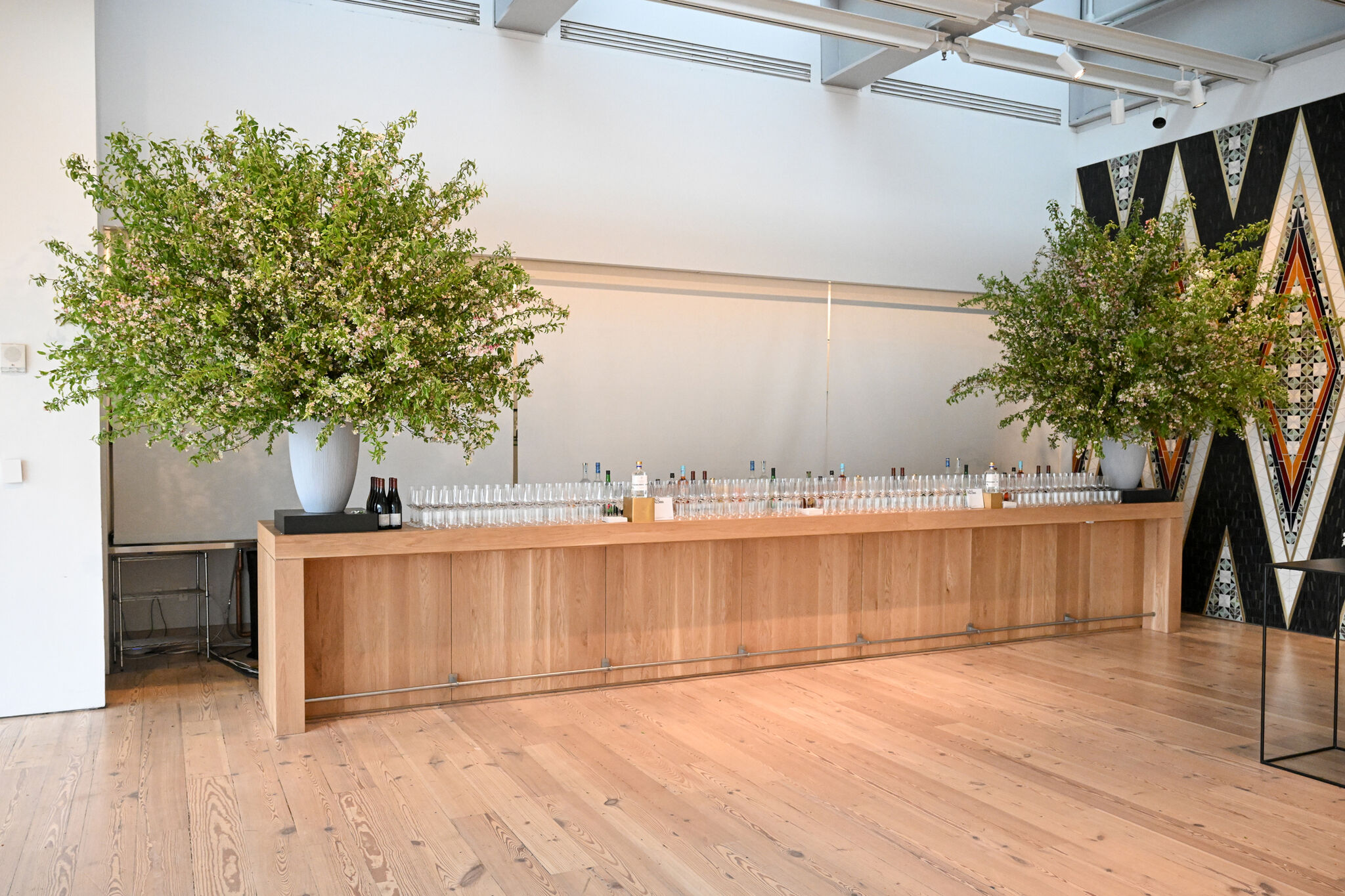 A long wooden bar with two large vases of greenery, lined with glasses and bottles, set in a spacious room with a patterned wall.