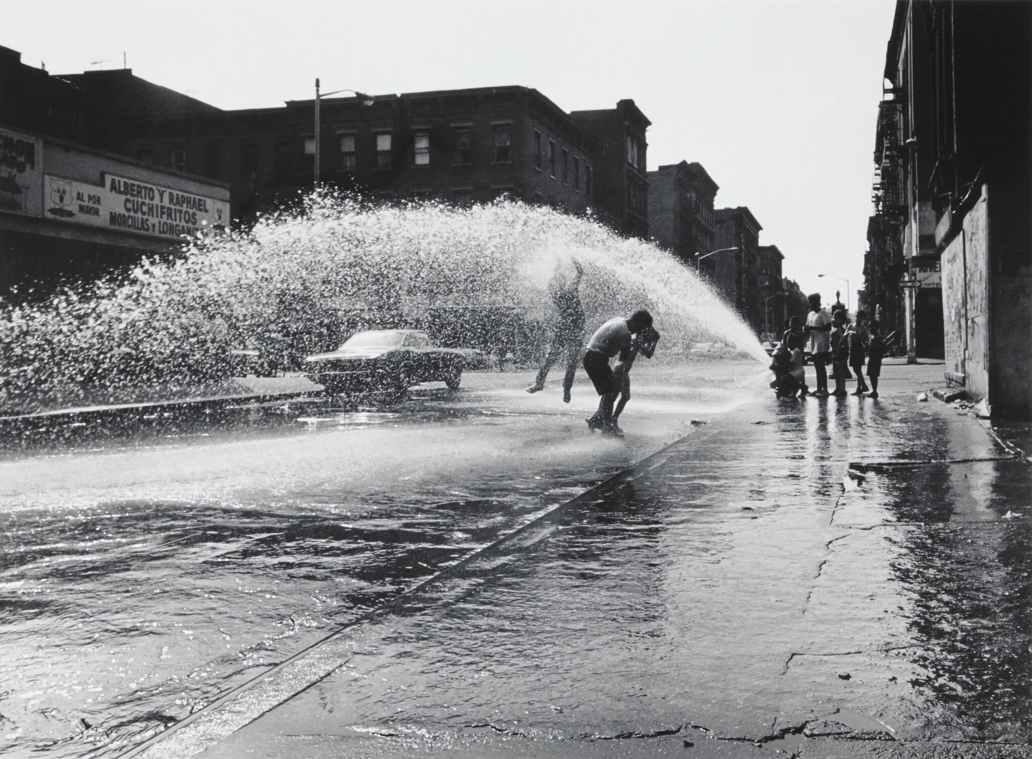 Children play in water spraying from an open fire hydrant on a city street, with buildings and a parked car in the background.