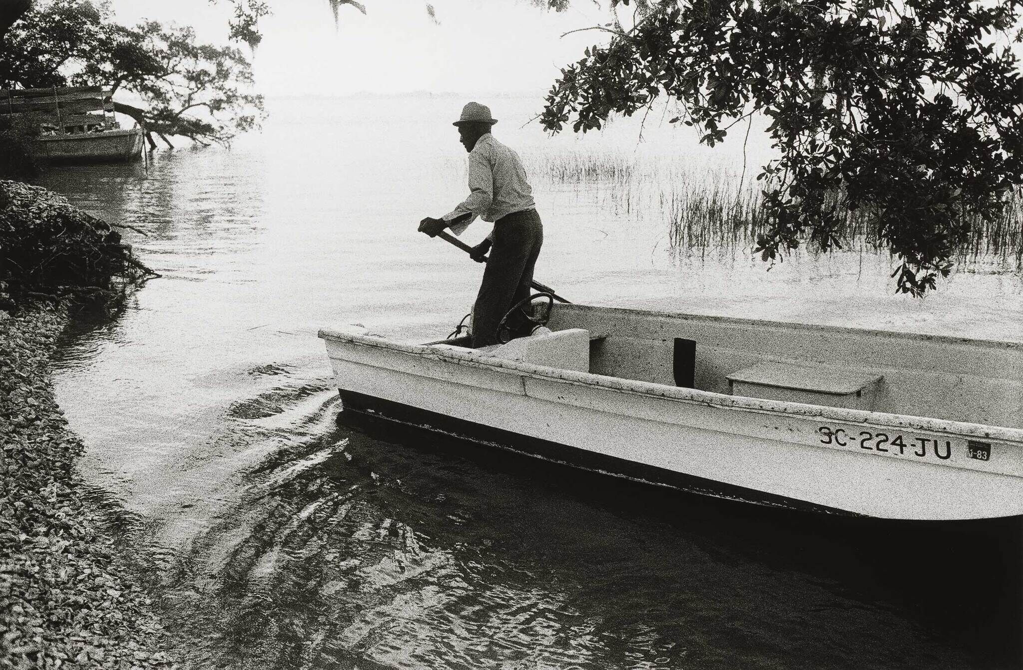 A man in a hat steers a small boat near a rocky shore, with trees and calm water in the background.