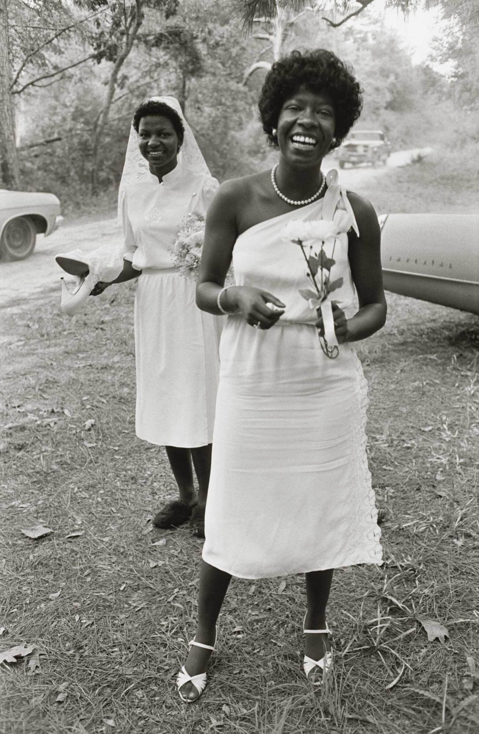 Two women in white dresses, one in a veil holding shoes, the other smiling with a flower, stand outdoors near a vintage car.