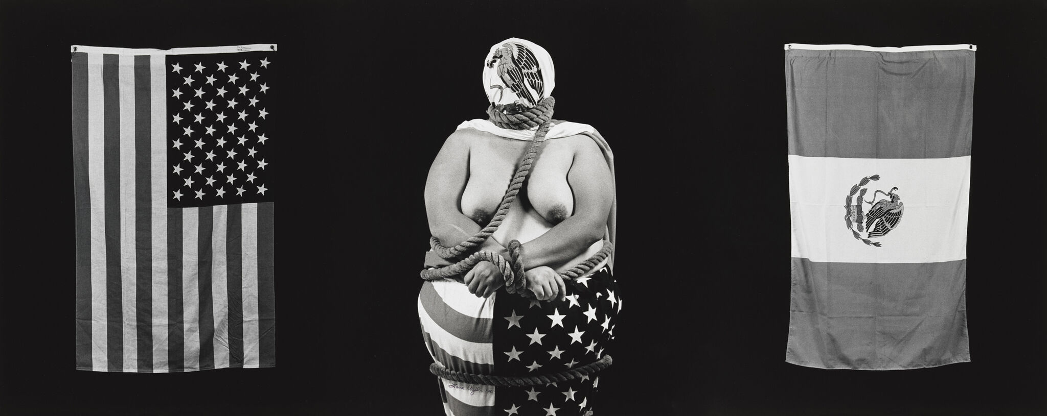 A person wrapped in a U.S. flag, bound with rope, and wearing a mask with an eagle design, flanked by U.S. and Mexican flags.