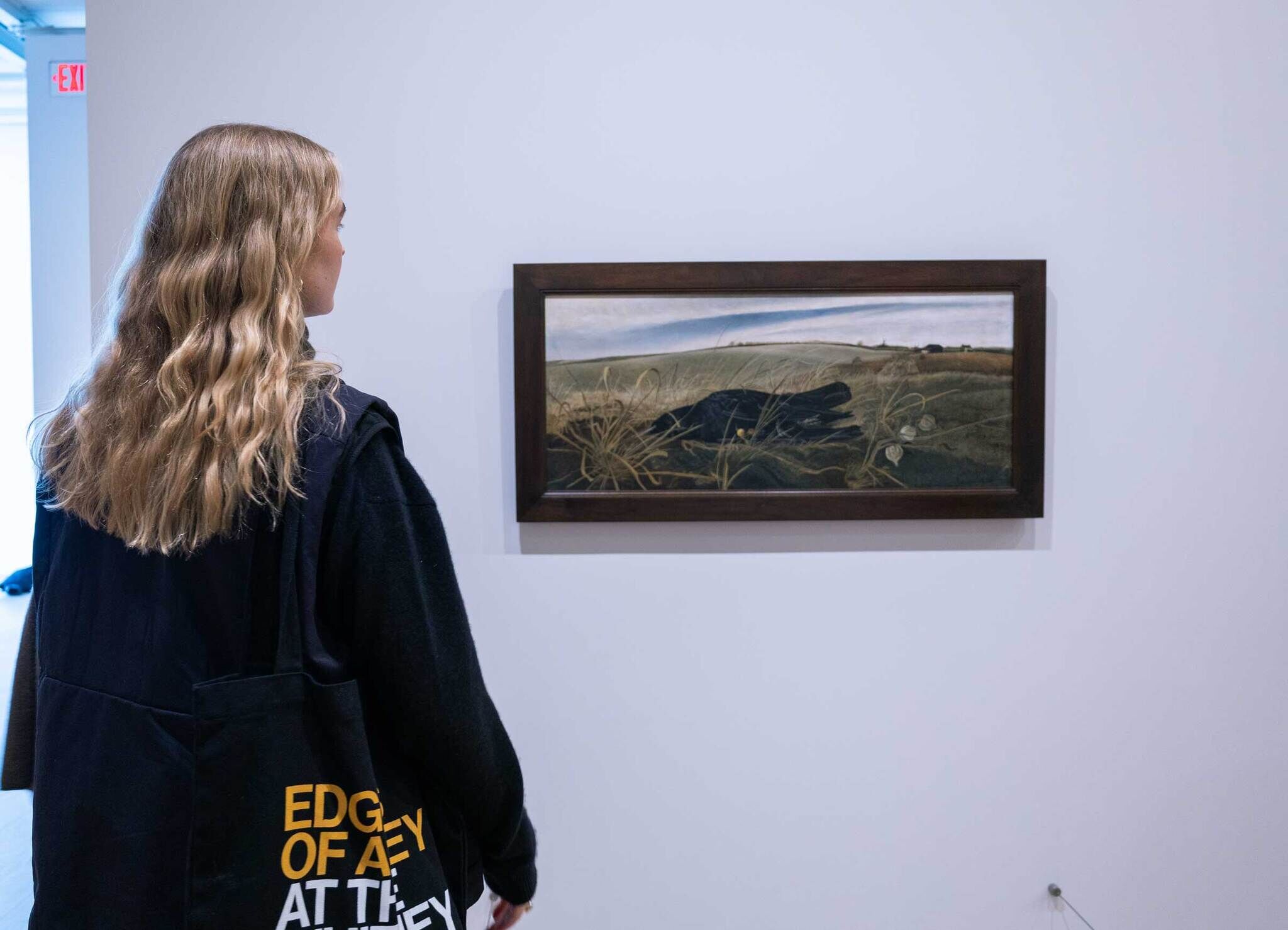 A person with long hair views a landscape painting in a gallery. They carry a black tote bag with yellow and white text.