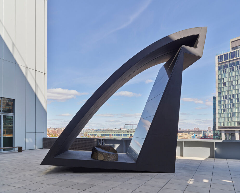  Modern outdoor sculpture resembling a stylized triangular portal, with city skyline and blue sky in the background.