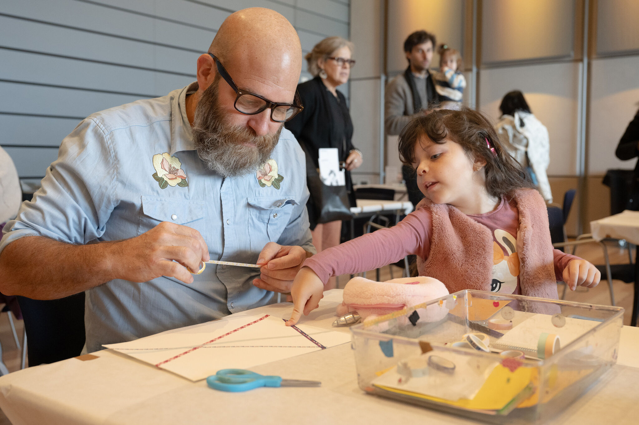 A man wearing glasses and a young girl are engaged in an art activity at a table during the Whitney Museum's Second Sunday event. The man is carefully applying tape to a sheet of paper while the girl, wearing a pink vest, points to something on the table, offering her input. Art supplies, including tape, scissors, and colorful materials, are spread out in front of them. In the background, other adults and children can be seen, adding to the atmosphere of a busy, creative, family-friendly event.