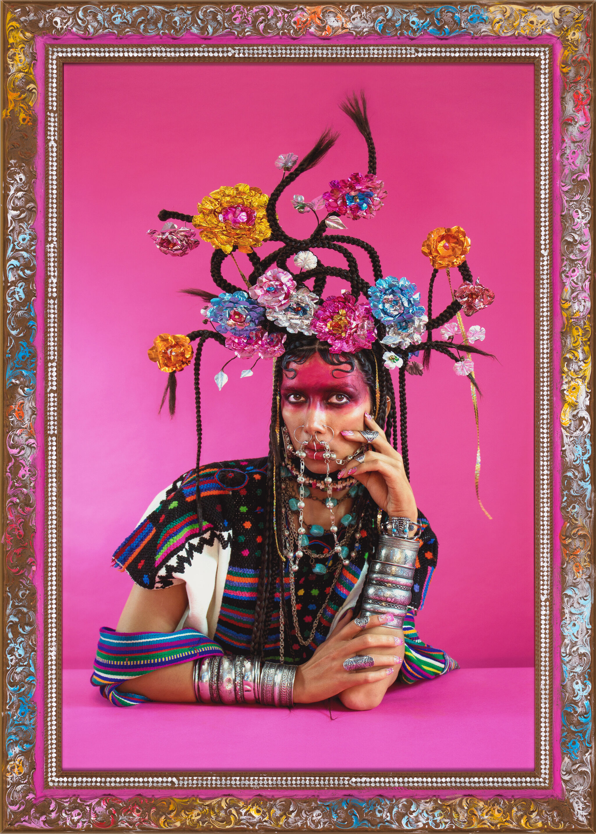 Person with vibrant makeup and elaborate floral headdress, wearing colorful attire and silver jewelry, set against a pink background in an ornate frame.