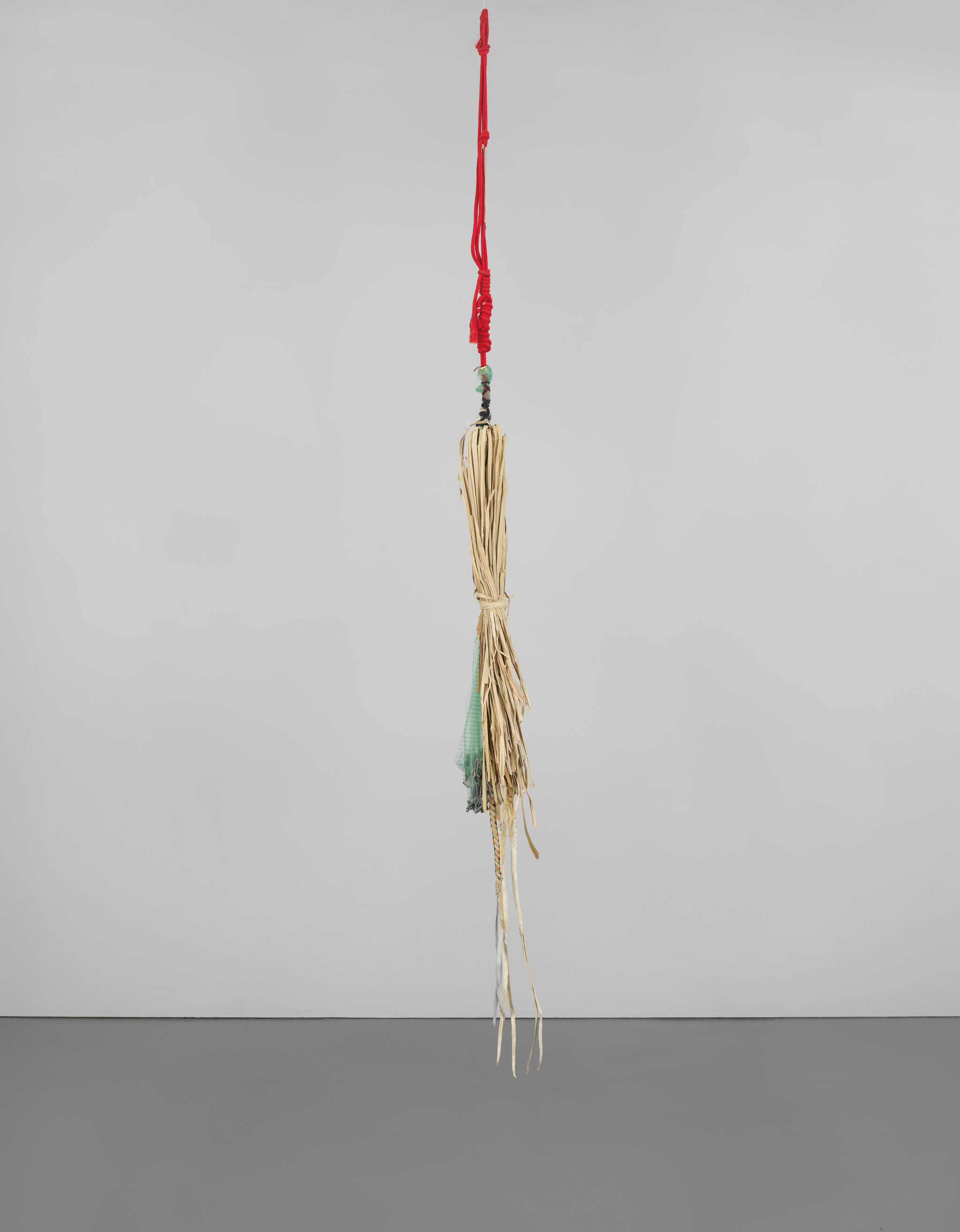 A hanging sculpture with a red cord, beige strands, and a green cloth, suspended against a plain gray background.