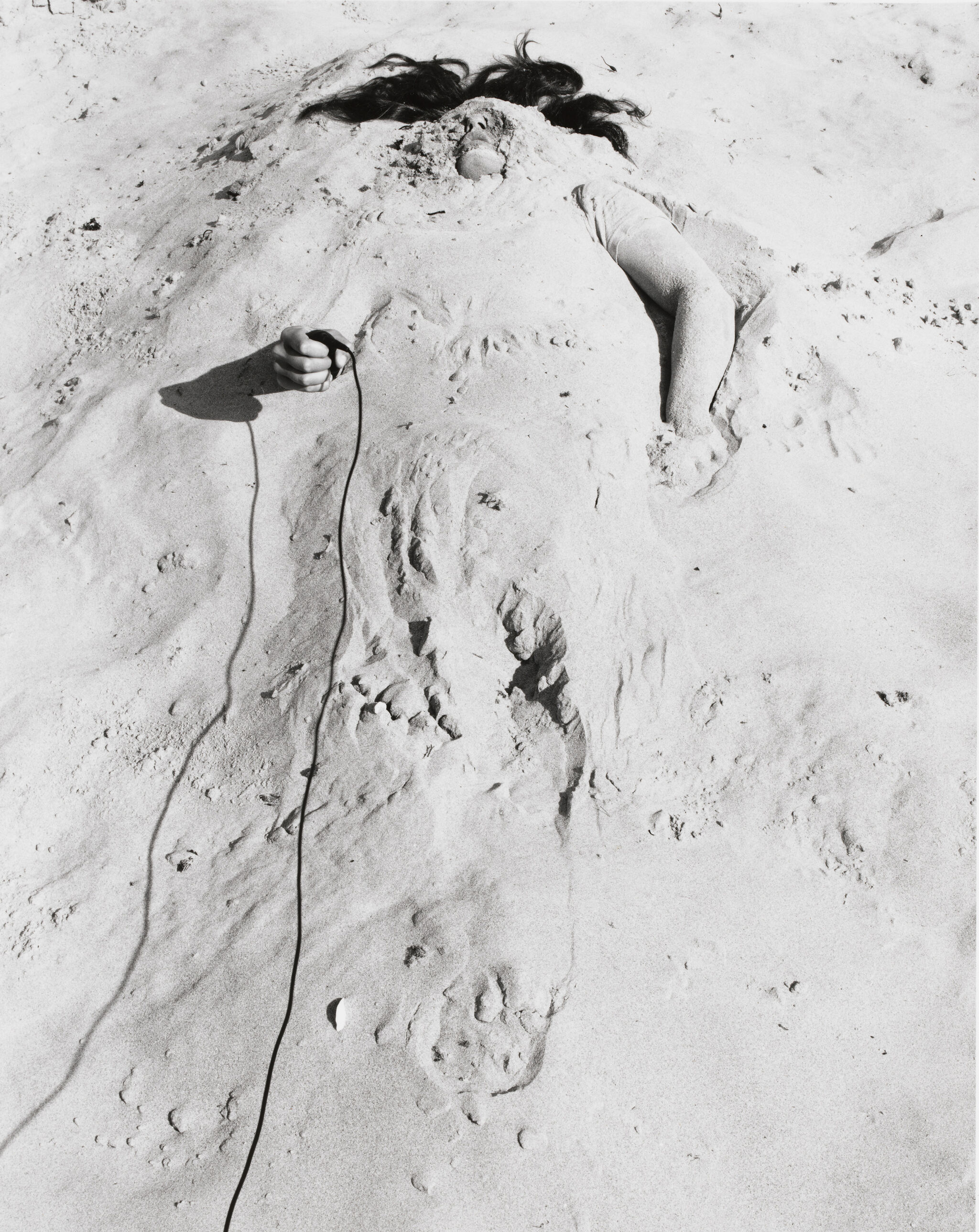 Person buried in sand with only hair and hand visible, holding a wire.