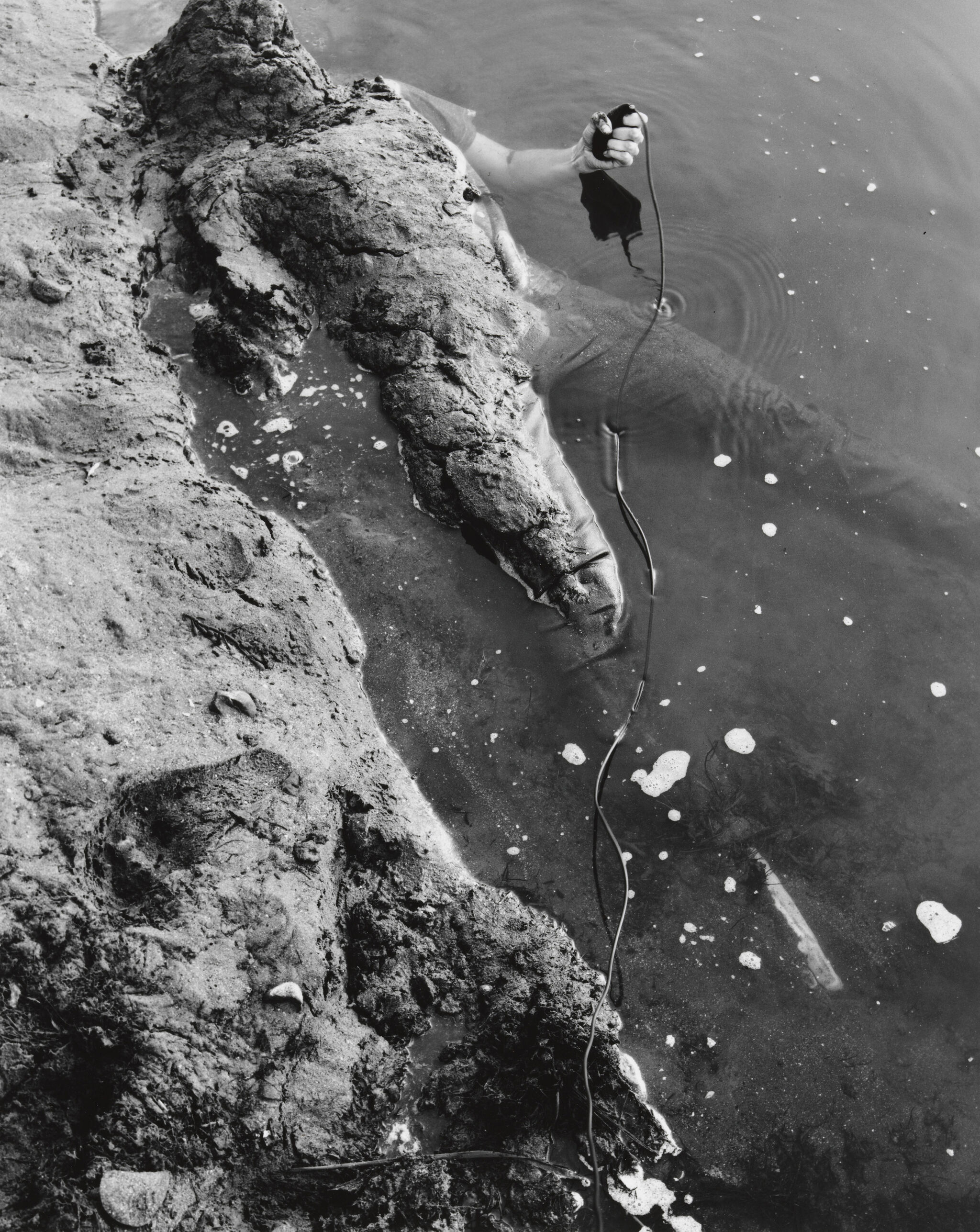 A hand holding a black object emerges from the water near a rocky shore, with a cable trailing into the water.