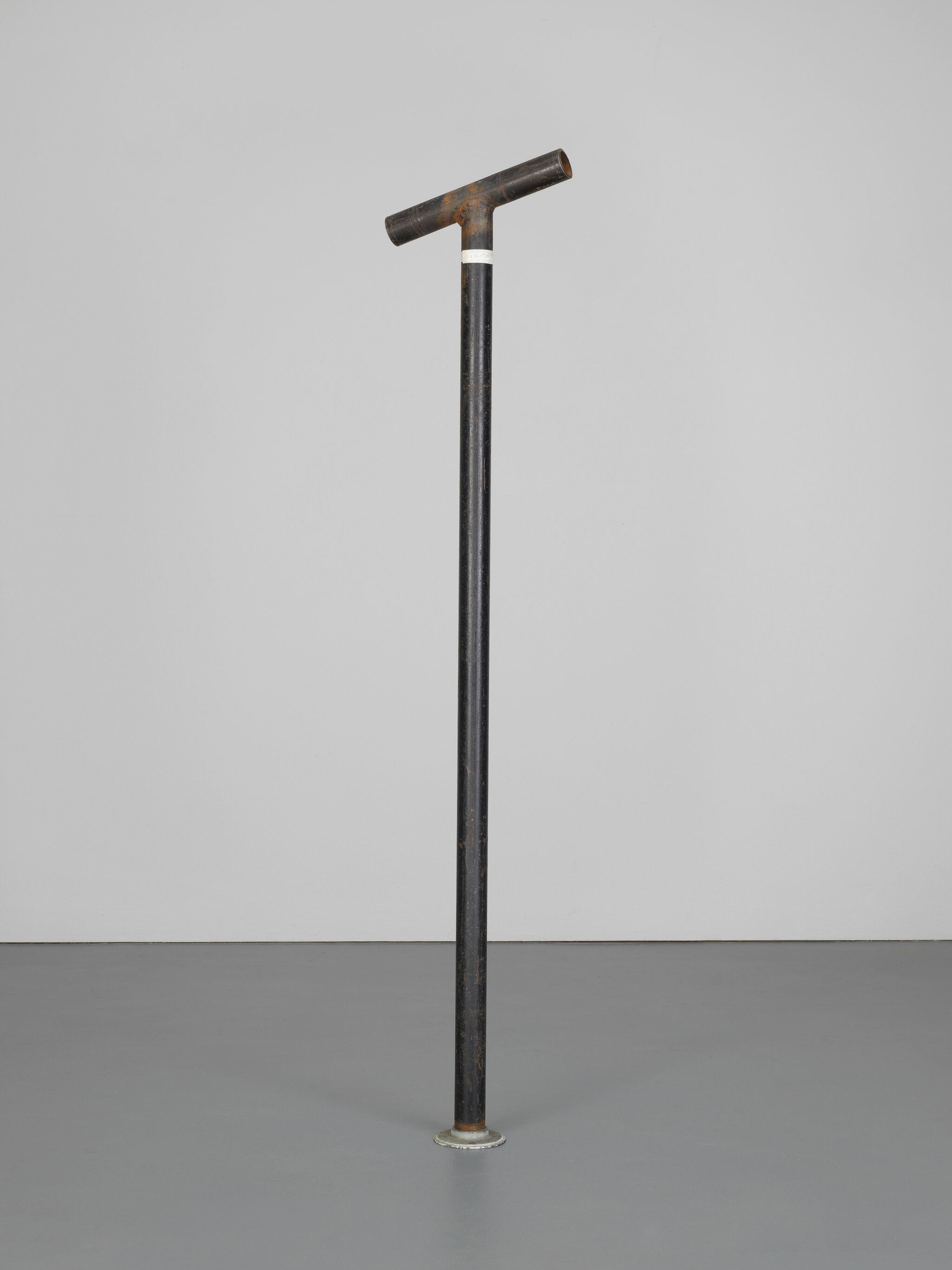 A tall, black metal pole with a T-shaped top stands upright on a gray floor against a light gray background.