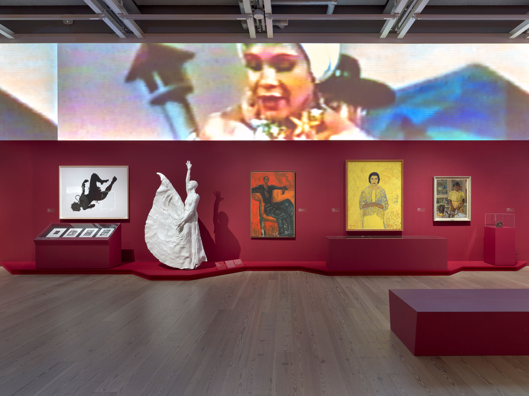 Art gallery with red walls, featuring paintings, a white sculpture of a dancer, and a large screen displaying a colorful image above.