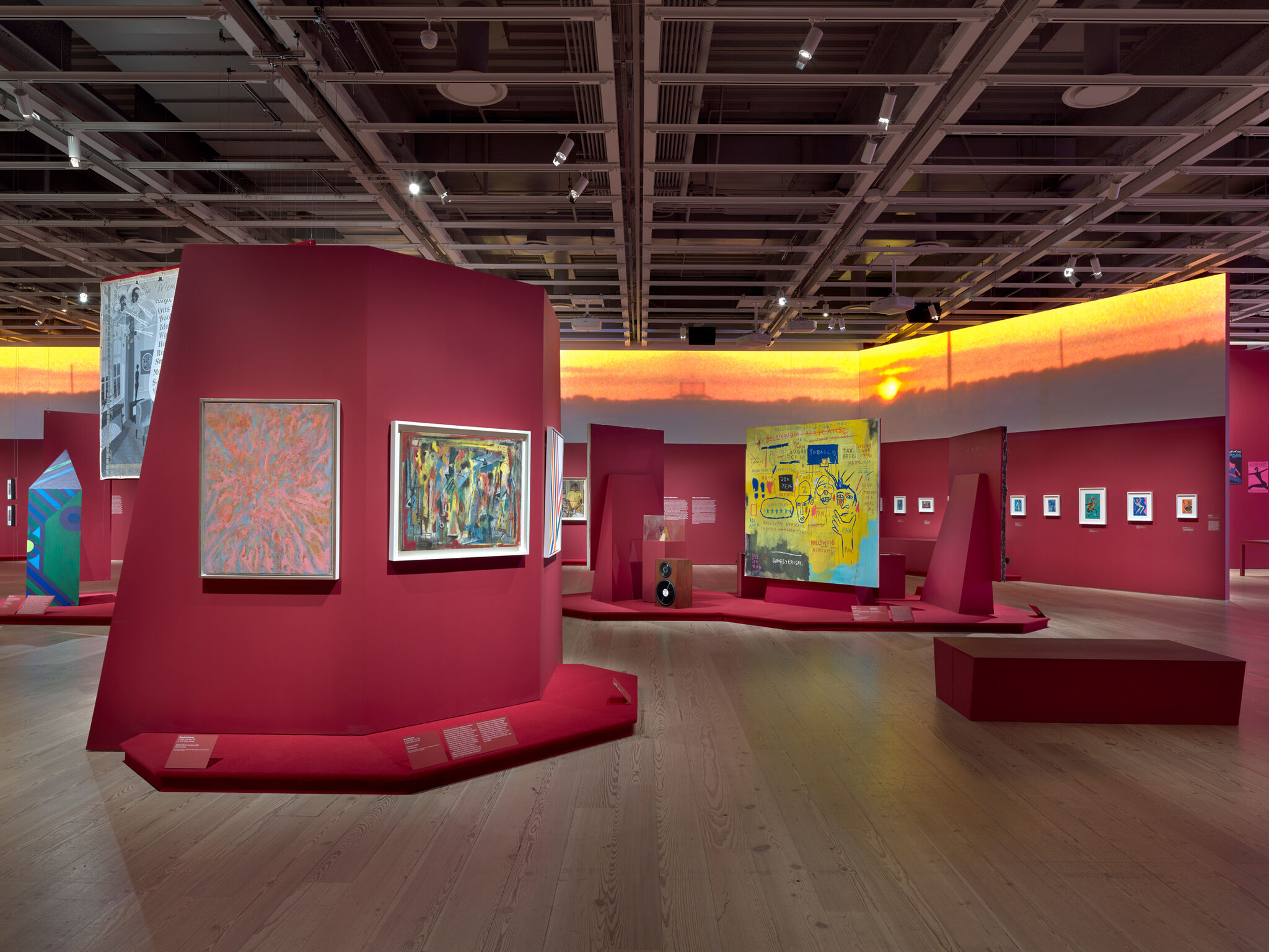 Art gallery with vibrant red walls, featuring various colorful paintings and installations. The ceiling has exposed beams and spotlights.